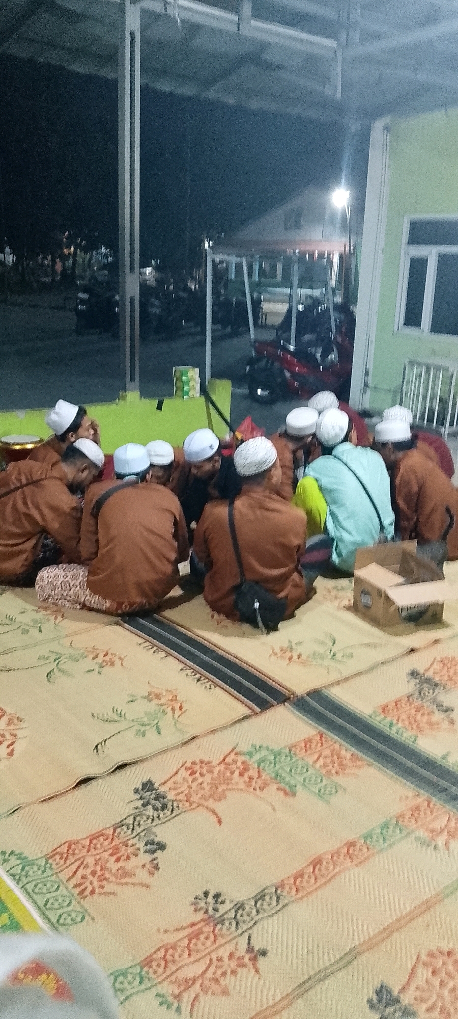 In Satriamekar, Indonesia, a group of at least ten men, all wearing white peci hats, are seated on the ground, facing inward and towards the center of their gathering. They are dressed in matching brown long-sleeved tunics and some are wearing patterned pants. The men are seated on woven mats that cover the ground, which are decorated with floral and geometric patterns in shades of cream, orange, and green.
Behind the seated men, the environment appears to be an open-air covered area, possibly a community hall or a mosque courtyard. Several motorcycles are visible parked in the background. A light pole with a bright light illuminates the scene from the right side of the image. A light green building with a white window frame is visible in the far right background. A cardboard box sits on the ground to the right of the group. The overall atmosphere suggests a communal gathering or religious activity taking place in the evening.