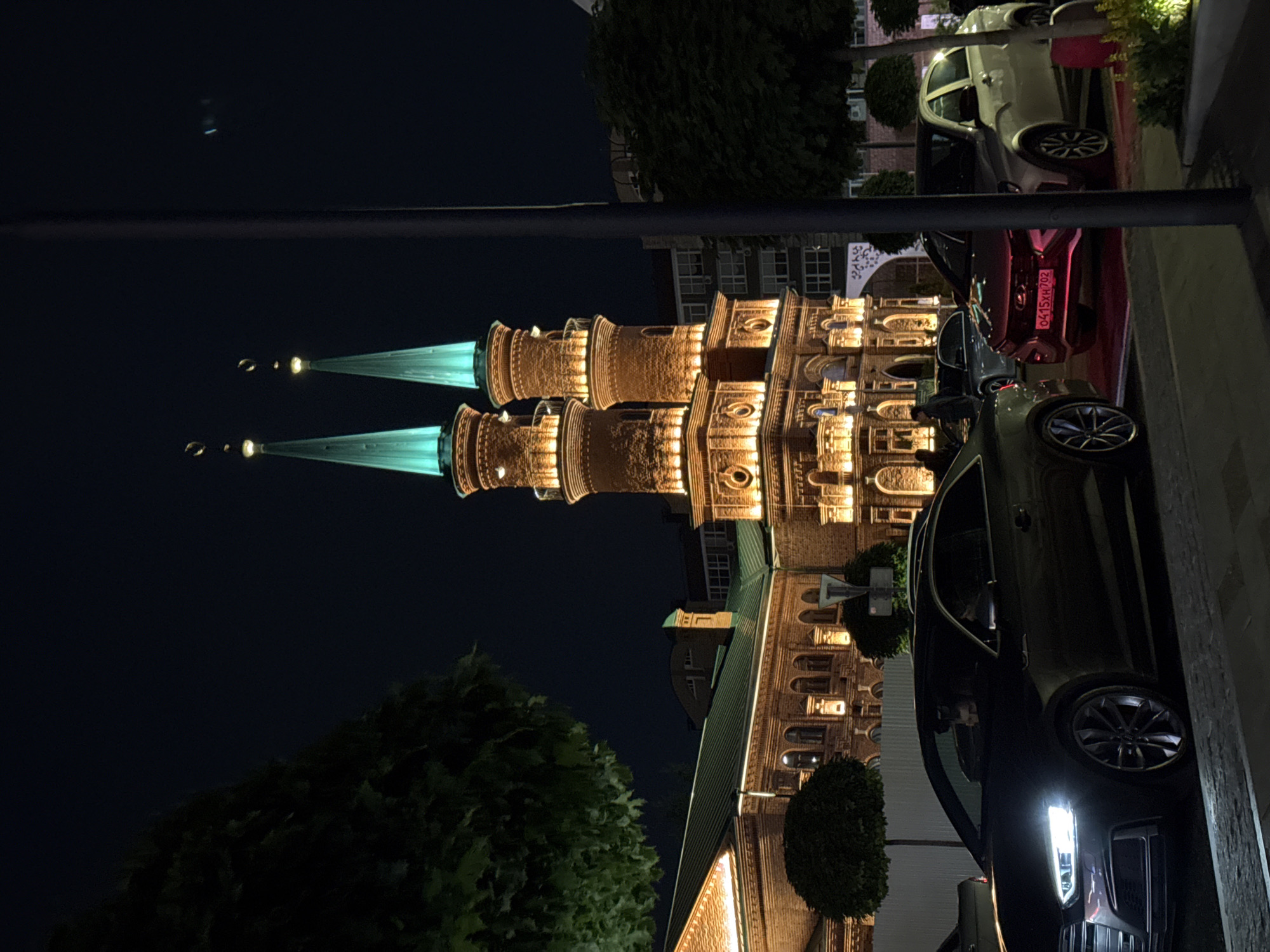 This is a night-time, eye-level, full shot of the Ufa Mosque in Ufa, Russia. The mosque features two tall, illuminated brick towers with teal-colored spires topped with crescent moons. The main building of the mosque is also lit with warm, golden lights and has a green roof. In the foreground, there are several parked cars. A dark-colored coupe is prominently positioned on the left, with its headlights on. To the right of the coupe, a red sedan is parked, and behind it, a silver hatchback. The street appears to be paved, and there are trees and a metal fence visible. The sky is dark blue, indicating nighttime.