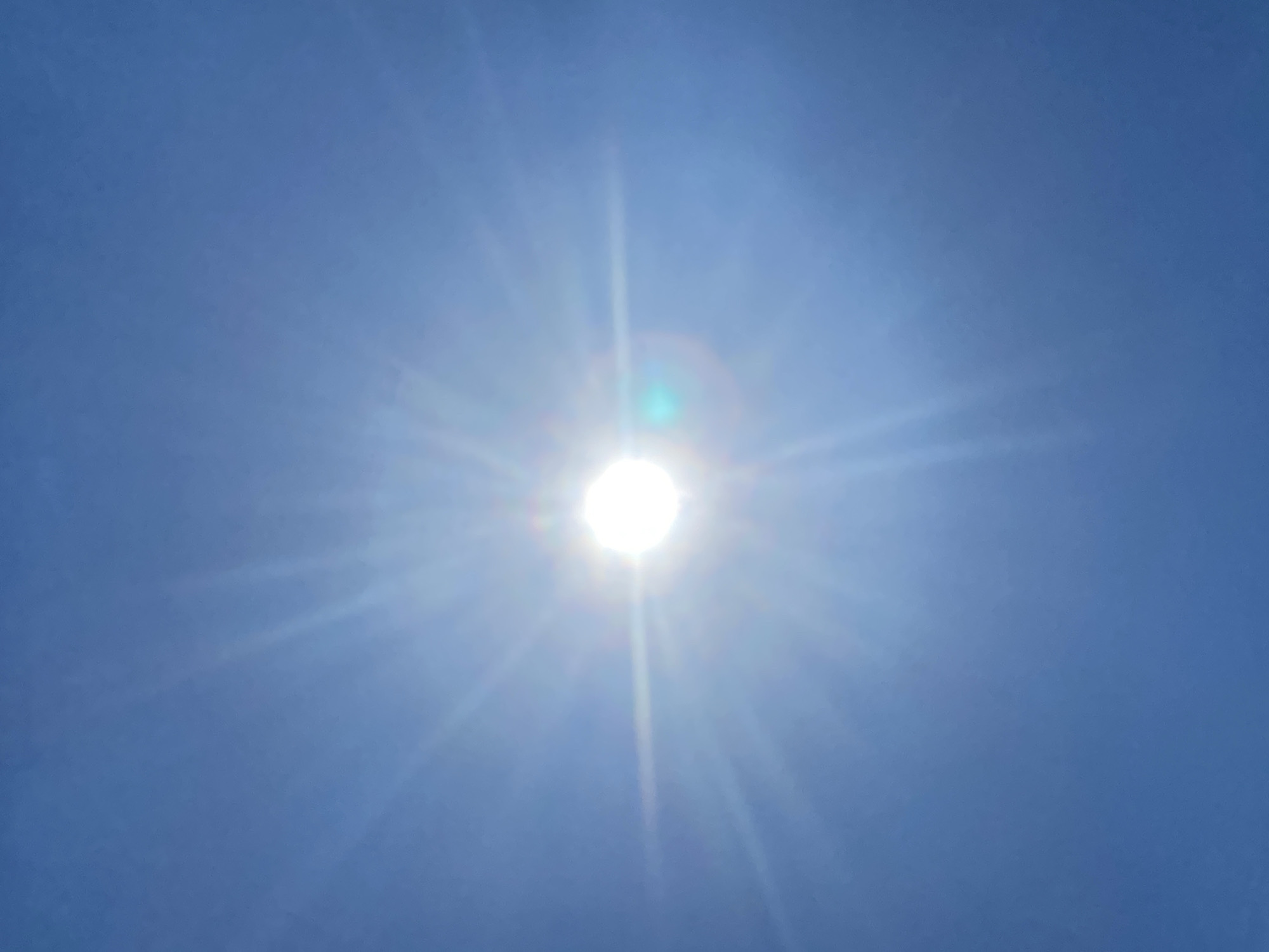 The image is a close-up, vertically oriented shot of the sun in a clear blue sky. The sun is the dominant feature, positioned slightly right of center, and appears as a bright, white disc. Sun flares radiate outwards from the sun, creating a starburst effect that fills much of the frame. The sky is a consistent, deep blue, with no clouds visible. There are some faint lens flares, including a noticeable green and blue circular flare to the right of the sun. The overall impression is of a very bright, sunny day. The location provided, Matiashivka, Ukraine, is an external environmental context for this image, but the image itself shows no discernible landmarks or features specific to any city or location.