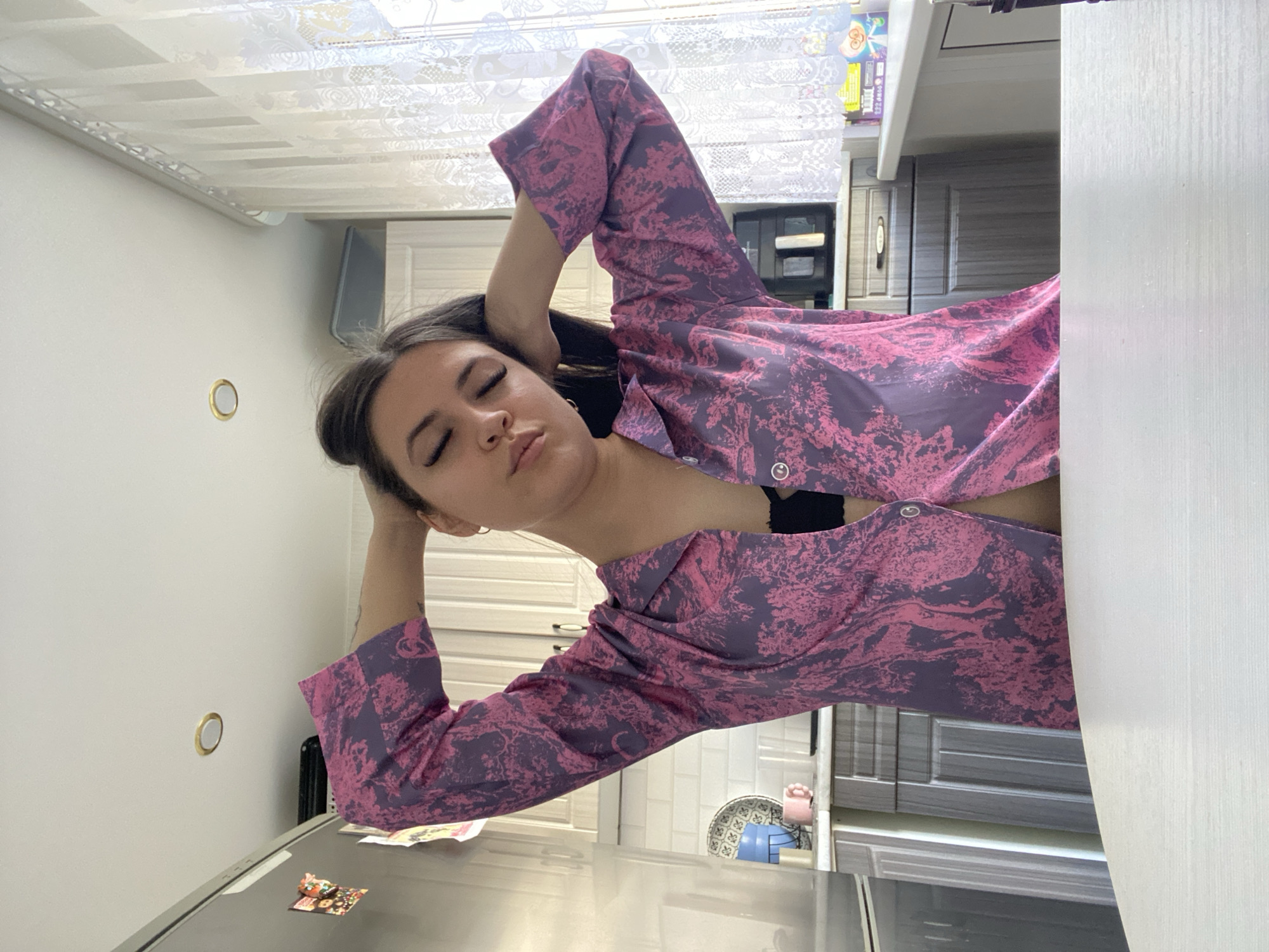 The image, taken in Chaikovsky, Russia, displays a young woman in a relaxed pose within a domestic kitchen setting. The photograph is rotated 90 degrees clockwise, orienting the woman horizontally across the frame.
The central subject is a young woman with long, dark hair. Her eyes are closed or mostly closed, suggesting a moment of rest or contemplation. She has her hands positioned behind her head, with her elbows pointing outwards, indicating a relaxed posture. She is wearing a long-sleeved shirt featuring a distinct pink and purple abstract or botanical pattern. A black bra or camisole is visible beneath her partially unbuttoned shirt. A small hoop earring can be seen on her left ear.
The background details suggest a kitchen. On the right side, a white cabinet or countertop section is visible. Behind the woman, a range of kitchen cabinets, varying in shades of grey and white with visible handles, line the wall. To the far left, a metallic surface, possibly the top of a refrigerator or a large appliance, is present. A circular gold-rimmed object, resembling a light switch or a decorative fixture, is mounted on a plain white wall. The ceiling features a textured or patterned finish. On a visible counter or shelf in the mid-ground, a blue bowl, a pink container, and some small packaged items are discernible. The overall scene conveys a calm, private moment in a home. No discernible text is visible anywhere in the image.