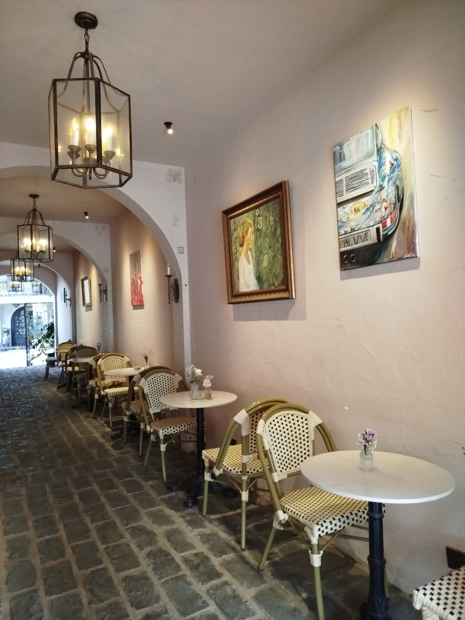 The image depicts an outdoor seating area of a cafe located in Phường Xuân Hương, Đà Lạt, Vietnam. The setting appears to be a walkway with arched openings, paved with grey cobblestones. Several small, round white tables with black bases are arranged along the walkway, each accompanied by wicker chairs with white and black patterned seats. Decorative elements include paintings on the wall and lanterns hanging from the ceiling. One painting shows a woman with blonde hair, and another depicts a stylized image of a vehicle. Small vases with single flowers are placed on some of the tables. The overall atmosphere is one of quiet outdoor dining.