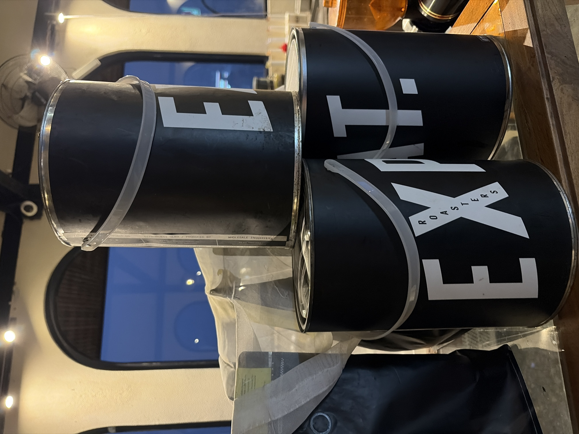 In Kintamani, Indonesia, three black cylindrical containers are stacked on a wooden surface. The top container, viewed from the side, displays a large white "E" and a partial "X." The middle container, also viewed from the side, shows a large white "P." The bottom container, in the foreground, features a large white "EX" and the word "ROASTERS" vertically arranged within the "X." The background shows arched windows, a ceiling fan, and bright lights. A light-colored fabric is draped over some of the containers.
