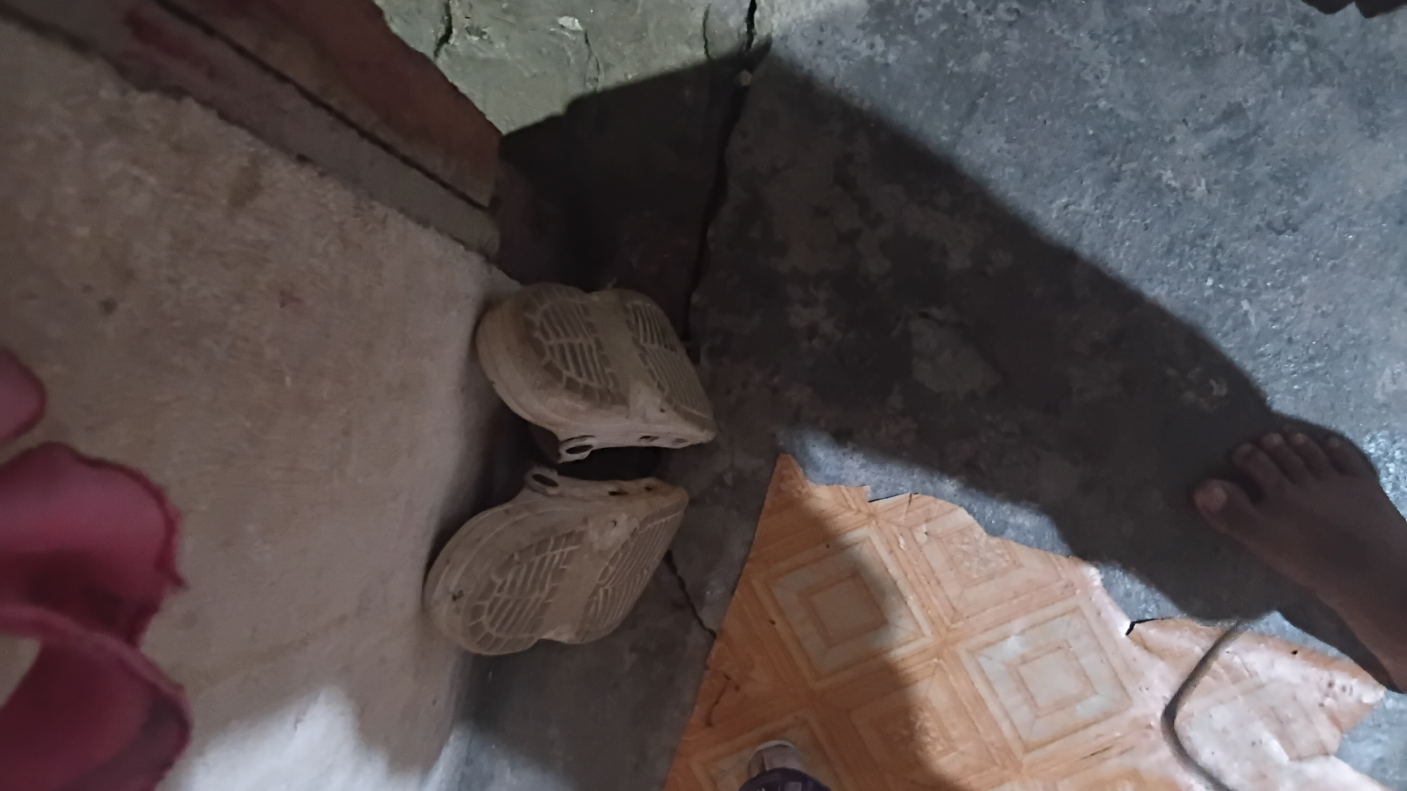 In a dimly lit room, two light-colored clogs are placed side-by-side on a beige rug. The clogs are facing away from the viewer, and their soles, with a distinctive pattern, are visible. To the left of the clogs, a shadow is cast on a cracked concrete floor, partially obscured by the clogs and the rug. A person's bare foot, with visible toes, is positioned in the upper right quadrant of the image, casting the large shadow. The floor also shows a section of orange and white tiled flooring in the upper right corner. The scene suggests a simple, indoor setting possibly in Prabumulih, Indonesia, where the clogs are resting.