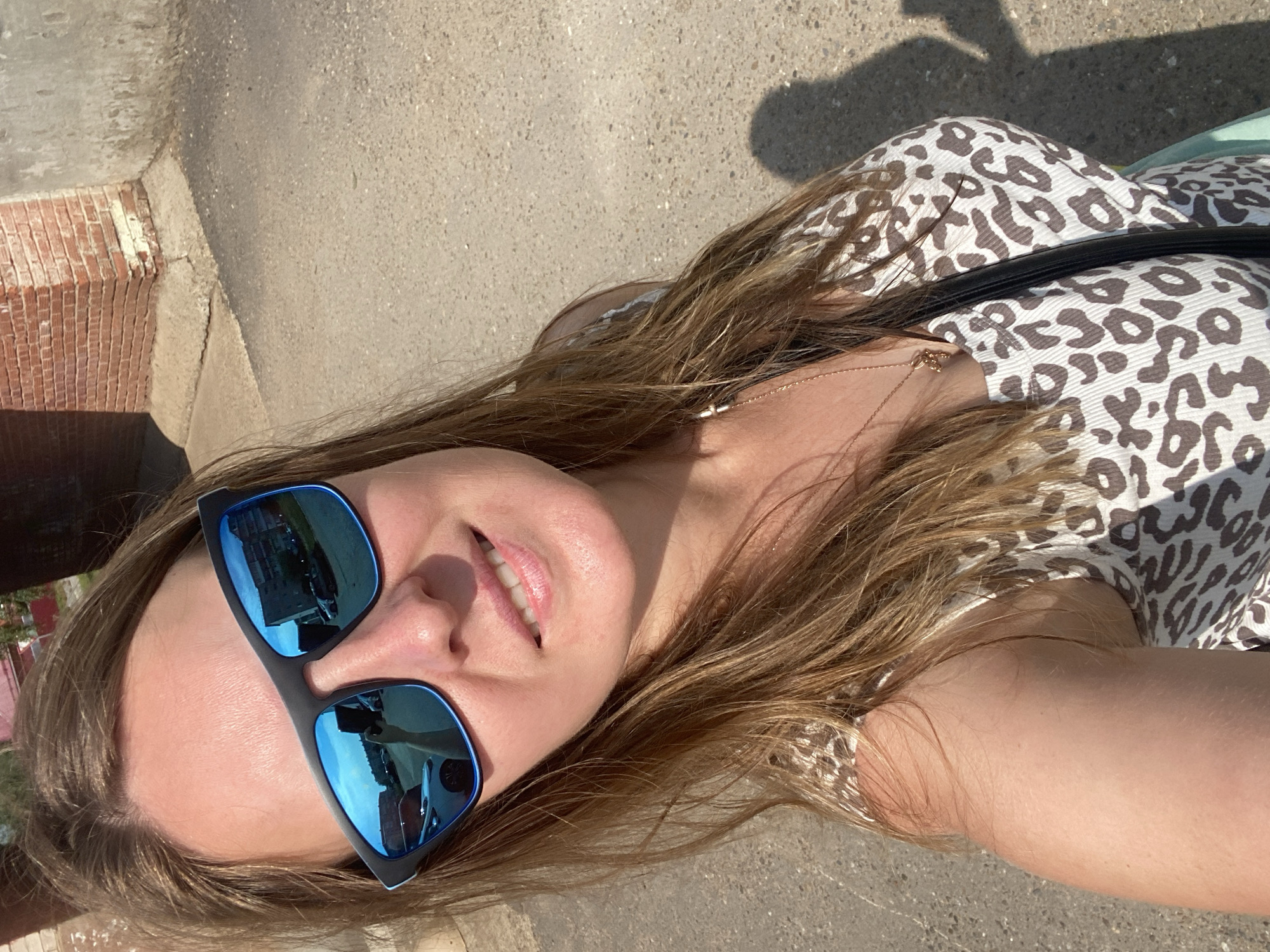 A woman is taking a selfie outdoors on a sunny day in Noviy, Russia. She is wearing sunglasses with blue lenses and a leopard-print top. The background shows a brick building and a concrete wall.
