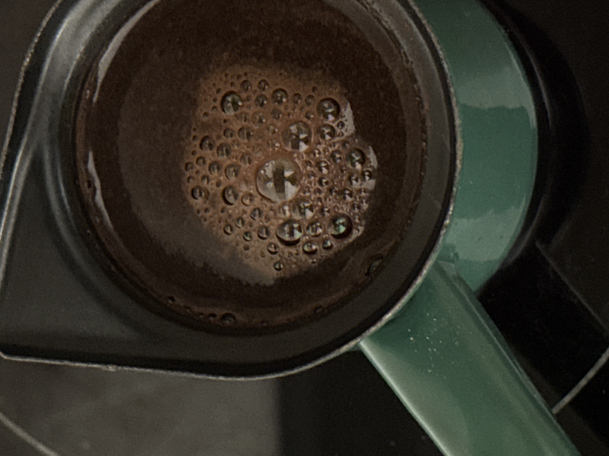 This close-up, overhead shot features a traditional coffee pot, commonly known as a cezve or ibrik, filled with freshly prepared coffee. The dark, rich brown liquid occupies the center of the frame, its surface largely covered with numerous small, light brown bubbles and a thicker layer of foam, indicative of a hot, freshly brewed beverage. Within the larger bubbles, distinct reflections, possibly from an overhead light fixture or a window, are visible as geometric patterns.
The pot itself has a dark, metallic rim, visible around the circumference of the coffee. Extending from the right side of the pot is a bright, deep green handle, which appears to be enameled or painted metal. The background is indistinct and dark, suggesting the pot is resting on a dark surface, possibly a stovetop or counter in an indoor setting.
Given the context of Budva, Montenegro, the image likely captures the preparation or presentation of traditional Balkan or Turkish coffee, a popular cultural beverage in the region. The scene conveys a simple, domestic moment focused on the appreciation of coffee. No people are visible, and no text is discernible within the image. The time of day and weather cannot be determined, but the focus on coffee suggests a morning or a break.