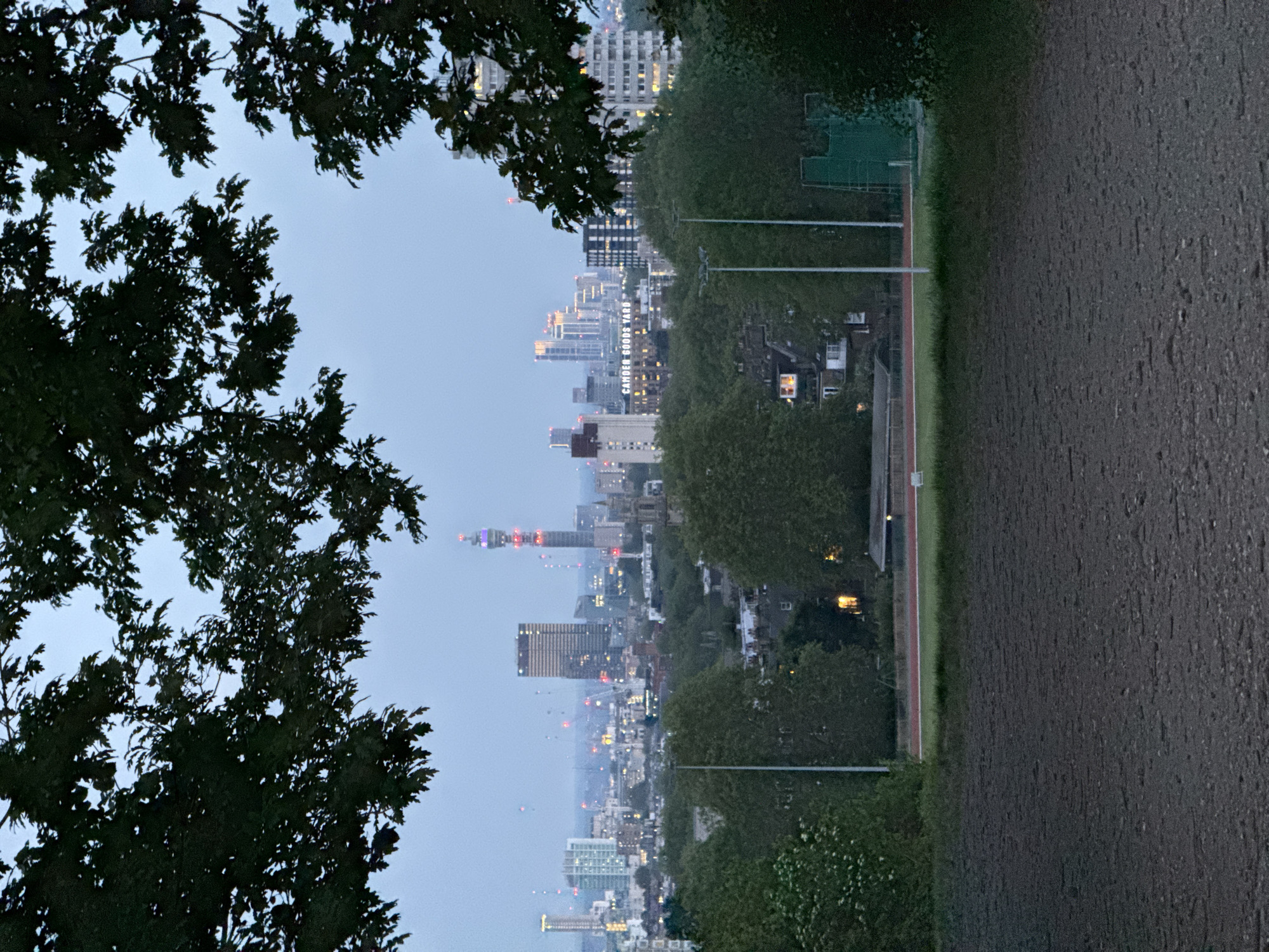 The image shows a view of the London skyline through tree branches. In the distance, several high-rise buildings are visible, illuminated with lights. The BT Tower is prominent with its distinctive red and white lights. Closer to the foreground, there's a running track and possibly a tennis court, bordered by trees and foliage. The foreground is dominated by a dark, gravelly or dirt area. The sky is overcast. The "Camden Goods Yard" building is recognizable in the middle distance.