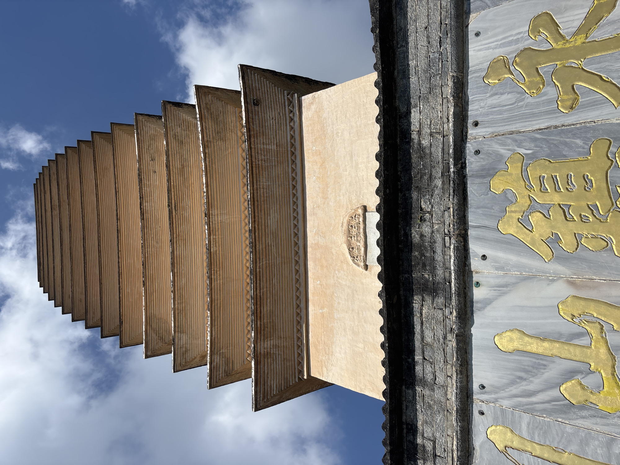 A low-angle, close-up outdoor shot captures the upper portion of the Three Pagodas of Chongsheng Temple in Dali City, China, against a bright blue sky with scattered white clouds. The pagodas, characterized by their tiered, light brown structures with receding eaves and ribbed detailing, dominate the frame. The foreground features a dark gray stone wall with a textured, wavy edge. Below this wall, a section of a light-colored marble plaque with large, ornate golden Chinese characters is visible, adding a decorative element to the scene. The image focuses on the architectural details of the pagodas and the decorative elements at their base, emphasizing their height and the interplay of light and shadow.