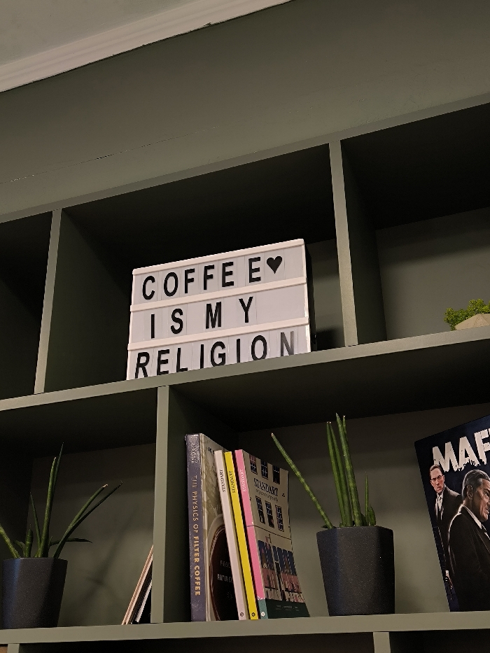 The image depicts an interior scene, specifically a section of a dark olive green, compartmentalized shelving unit in Vinnytsia, Ukraine.
On the upper shelf, a white light-up letter board is centered within a cubby, displaying the black text "COFFEE IS MY RELIGION" with a black heart symbol positioned after "COFFEE." To the right, within an adjacent cubby, a small, light-colored object topped with green moss or artificial greenery is visible.
The lower shelf contains various items arranged within its cubbies. In the leftmost visible cubby, a small, dark grey hexagonal planter holds a green Sansevieria cylindrica (African spear plant). A stack of several books with diverse spines (including purple, yellow, and pink) stands upright in the central lower cubby. A prominent title visible on one book is "THE PHYSICS OF FILTER COFFEE," and another is "STANDART." To the right, another identical dark grey hexagonal planter contains a similar Sansevieria cylindrica plant. Leaning against the far right side of the cubby is a dark-covered magazine or book featuring the word "MAFIA" at the top right, with two men in suits depicted; one appears to be actor Robert De Niro. No active people are present in the immediate scene.