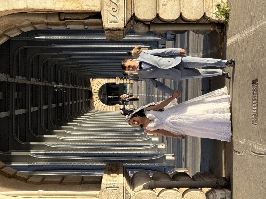 A wedding couple walks hand-in-hand along the pedestrian walkway of the Pont de Bir-Hakeim bridge in Paris, France. The female subject, positioned on the left, wears a white, floor-length wedding gown with lace details, a veil, and a tiara. Her left eye exhibits a slight squint. The male subject, on the right, wears a light gray three-piece suit, a white shirt, and a black bow tie. Both individuals appear to be of East Asian descent. They walk forward, with the female holding the male's right hand with her left, and the male holding the female's left hand with his right.
The environment consists of the bridge's architectural structure, featuring repetitive metal arched supports creating a long, tunnel-like passage. Stone piers with carved embellishments flank the foreground. The ground is a paved walkway with visible cracks and a rectangular metal grate. Strong natural light from the right side illuminates the subjects and bridge structure, casting defined shadows. In the distant background, several blurred figures of other pedestrians are visible, and an archway reveals an urban street with hints of traffic signals beyond.