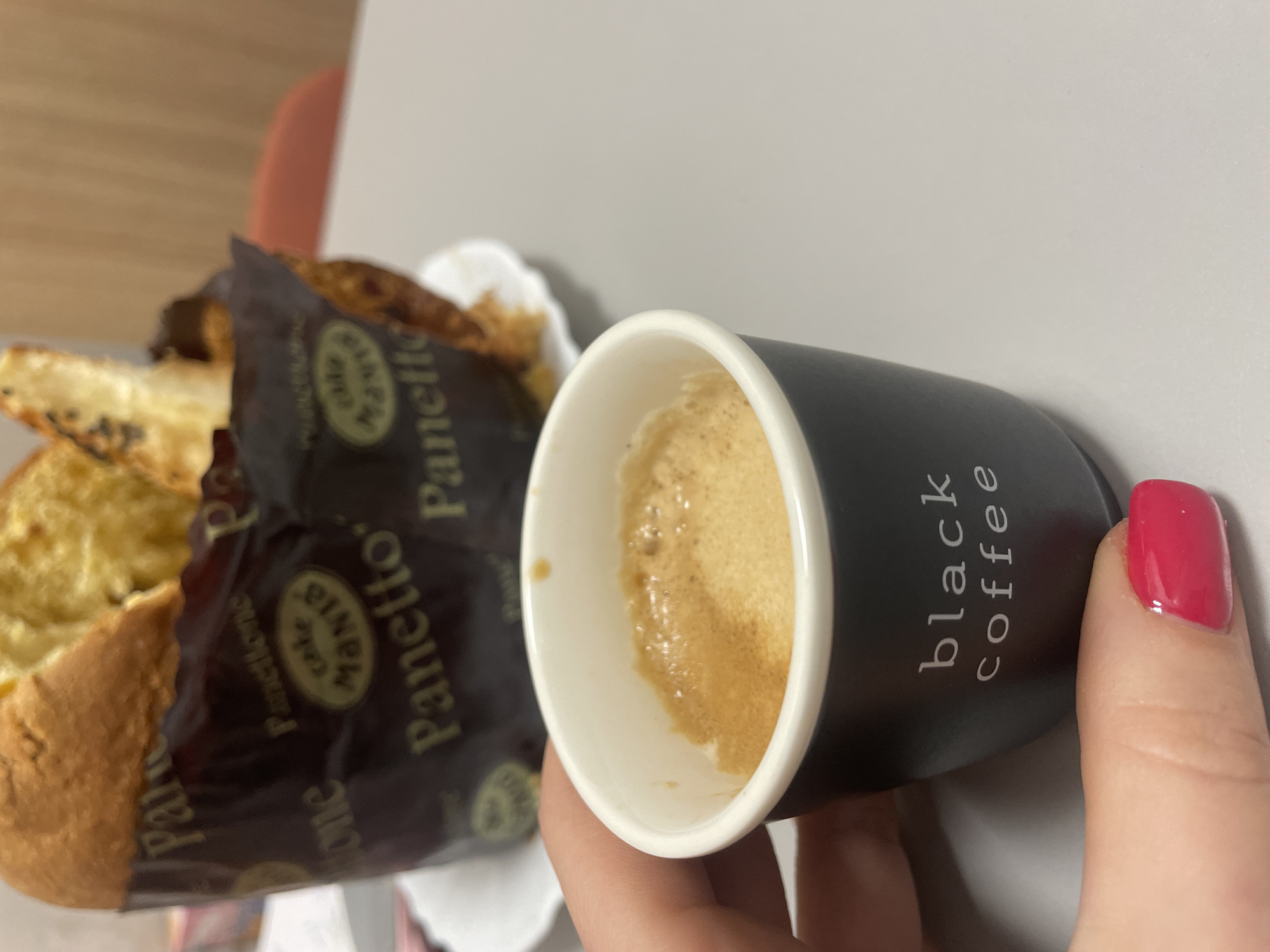 A partially consumed cup of black coffee, held in a person's hand, is positioned in front of pieces of Panettone, likely cake, in a Cafe Manza Panetto wrapper. The location is possibly Sofia, Bulgaria, based on the Panetto branding. The coffee cup is black with "black coffee" printed in white lettering. The person holding the cup has a pink nail polish on their nail. The background consists of a light-colored surface and a hint of an orange chair or wall.