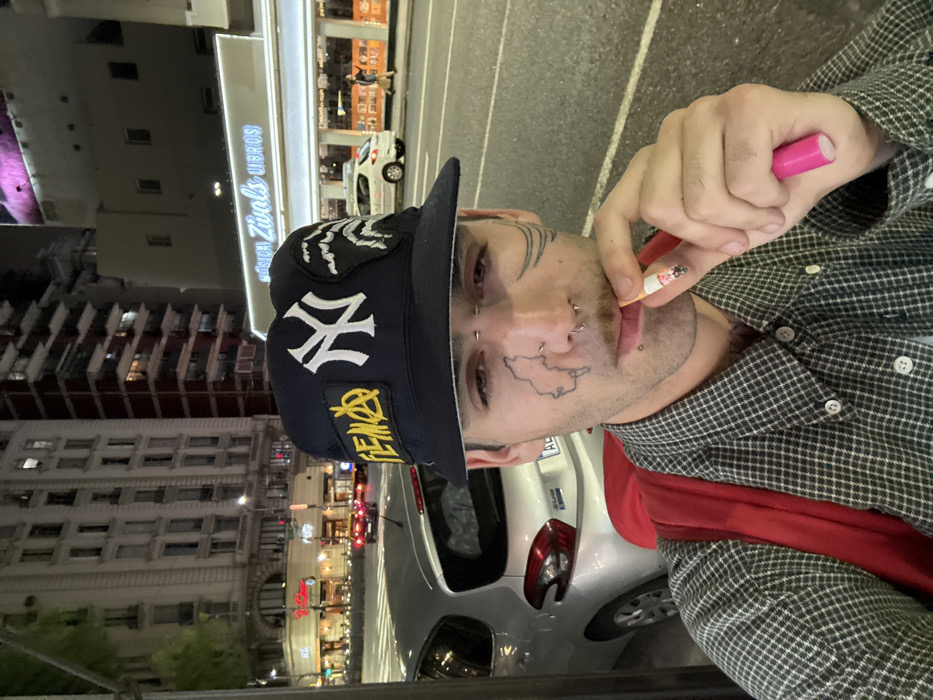 An image taken at night depicts a young adult male in the foreground, positioned on an urban street in Buenos Aires, Argentina, specifically near Avenida Corrientes as indicated by the prominent "MÚSICA ZIVALS LIBROS" sign in the background. The male subject has dark hair, a mustache, and multiple facial piercings including a bridge piercing, a spiked septum ring, a nostril stud, a philtrum stud, and a lower lip ring. He exhibits facial tattoos: a map-like outline on his left cheek and stylized lines extending from the outer corner of his right eye. He wears a dark blue bucket hat featuring a white New York Yankees logo, a yellow patch with "PEMA" or "FEMA" style text incorporating an anarchy symbol, and a black and white wave-like or tribal design patch. His attire includes a small-checked plaid button-up shirt and a bright red backpack. He holds a lit cigarette with an orange filter between his lips, gripped by his right hand, which also holds a distinct pink cylindrical object, possibly a vape pen or lighter, beneath the cigarette.
The background shows a street illuminated by artificial light sources. A silver Peugeot 208 hatchback is parked to the left of the subject. Across the street, the well-lit facade of "MÚSICA ZIVALS LIBROS" is visible, with several pedestrians on the sidewalk. Beyond Zivals, a white car is parked. Further down the street, a multi-story classical-style building with numerous illuminated windows is prominent on the left, alongside other urban structures. The street appears to have multiple lanes, and reflections on the pavement suggest damp conditions. Vehicle taillights are visible in the distance.