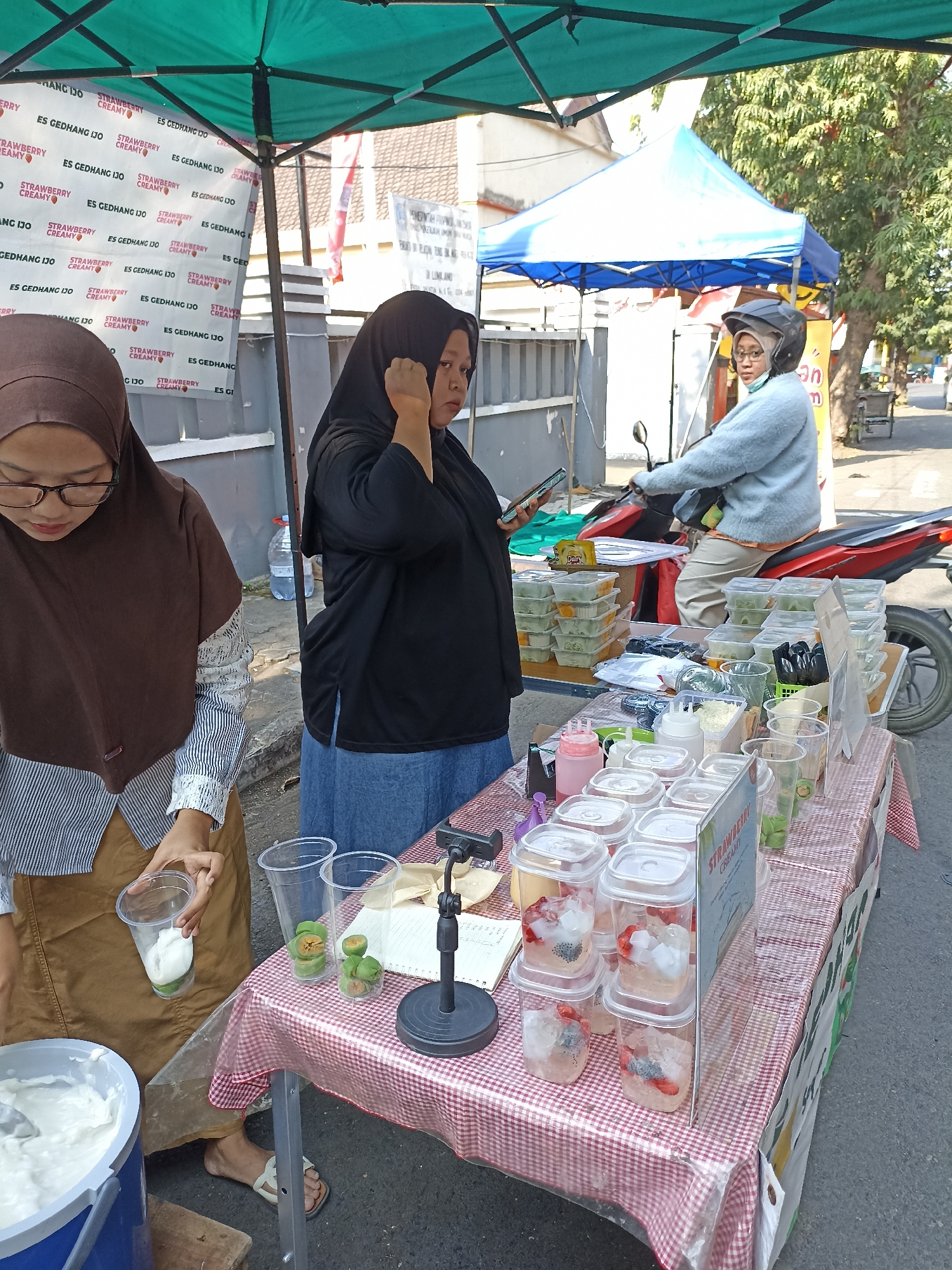 This image captures an outdoor scene in Lumajang, Indonesia, likely a street-side vendor stall. Two women are present. One woman, wearing a brown hijab and glasses, is actively preparing a drink, holding a clear plastic cup filled with a white creamy substance and a green slice. She is dressed in a striped long-sleeved shirt and a brown skirt. The second woman, wearing a black hijab and a blue skirt, stands behind the table, looking at her phone. She is positioned near a display of various drinks in clear plastic containers.
The stall itself is covered by a teal canopy and features a red and white checkered tablecloth. On the table, there are numerous containers of drinks, some filled with ice, fruit pieces, and seeds. There are also small containers of ingredients and bottles of sauces or syrups. In the background, a person wearing a helmet is seated on a red motorcycle. The environment appears to be a sunny street, with trees and buildings visible in the distance. A sign on the stall has text that is partially visible, including "STRAWBERRY CREAMY" and "ES GEDHANG IJO". A water bottle and a white bucket containing a white substance are also present near the stall.