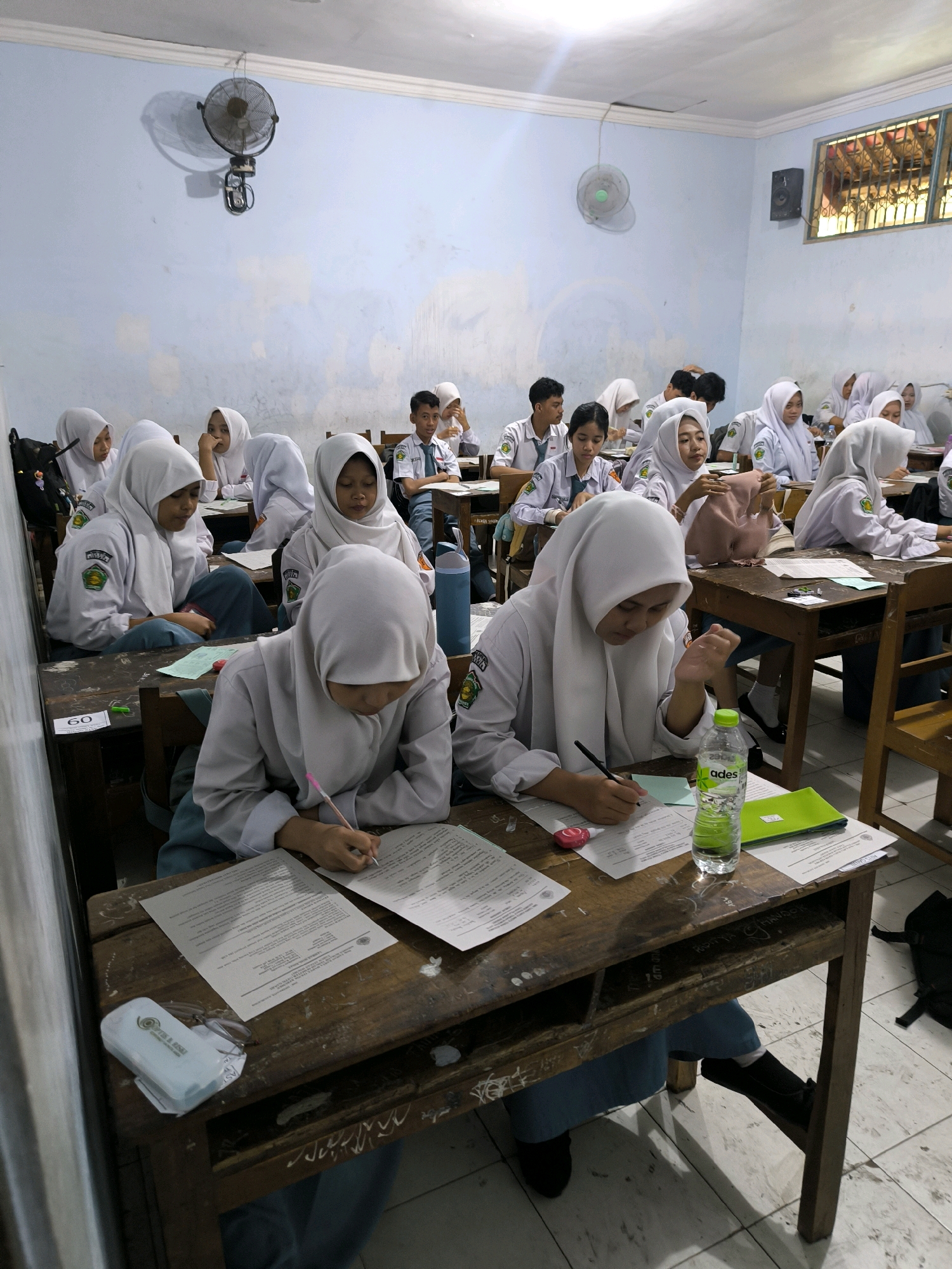 A classroom scene in Kebumen, Indonesia, captures several female students wearing white hijabs and school uniforms, likely during an examination or lesson. The students are seated at wooden desks, focused on their papers. One student in the foreground is actively writing with a pink pen. A bottle of "ades" mineral water is visible on her desk, along with other papers and a green folder. The classroom appears somewhat aged, with a fan mounted on the light blue wall and a window with bars. The overall atmosphere suggests a typical educational setting.