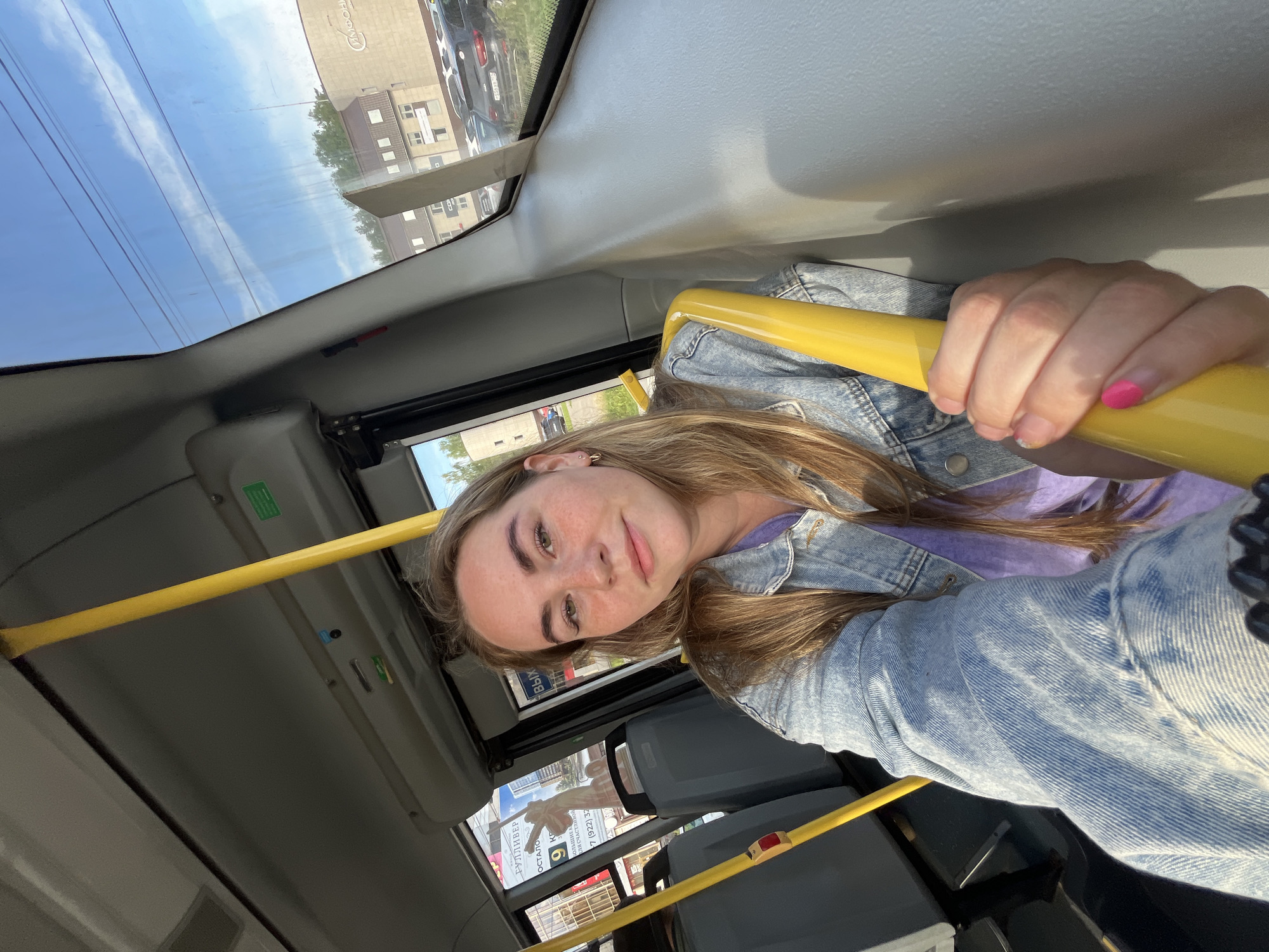 A young woman is captured in a selfie taken inside a public transport vehicle, likely a bus or trolleybus, during daytime in Chaikovsky, Russia. She has long, light brown hair and freckles, wearing a denim jacket over a purple top. She looks calmly towards the camera with a neutral expression or slight smile. Her right hand, with bright pink nail polish on one finger, grips a yellow horizontal grab rail. The vehicle's interior features light gray panels on the ceiling and upper walls, and dark gray seating.
Bright sunlight streams through the windows, illuminating a clear blue sky with wispy clouds and overhead power lines. The urban landscape outside includes multi-story buildings, parked cars, and visible greenery. Several signs with Cyrillic text are partially visible, including "РУЛЛИВЕР" (RULLIVER), "СИМФОНИ" (SYMPHONY), "Я СЧАСТЛИВОЙ" (I AM HAPPY), and a partial phone number "7 (922) 32", confirming the Russian location. The image conveys a sense of a relaxed journey through the city.