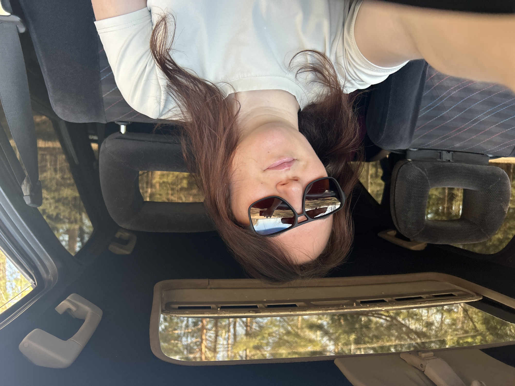 A person wearing sunglasses is inside a car with a sunroof. The seat has a gray headrest and striped fabric. The view through the car windows and sunroof shows trees. The location is not confirmed as Noviy, Russia.