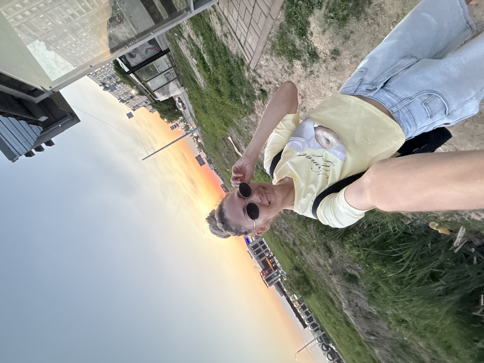 A woman wearing sunglasses and a yellow t-shirt is posing for a selfie. The woman is standing on a path next to a grassy area. In the background, there is a sunset and buildings. This location is in Заволжье, Russia, specifically near a bus stop with a glass structure. The woman is wearing light-wash jeans and a black bag. Her hair is styled in a bun.