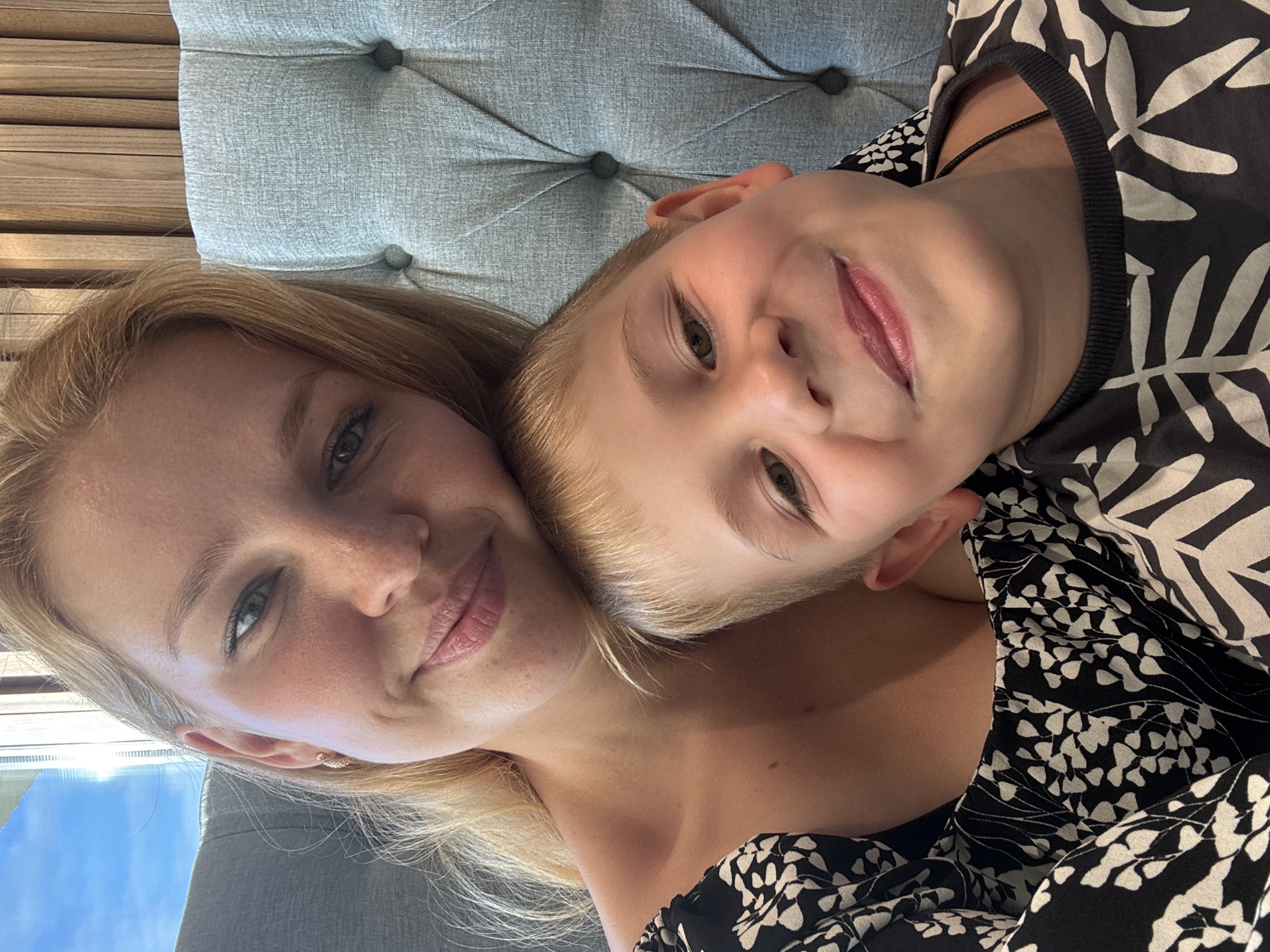 A close-up, selfie-style photograph shows a blonde woman and a young boy. The woman is on the left, her head tilted slightly, with her blonde hair falling over her shoulders. She has light skin, light-colored eyes, and is smiling. She is wearing a black and white floral patterned top. The boy is on the right, resting his head on the woman's shoulder. He has blonde hair and is smiling broadly, showing his teeth. He is wearing a dark shirt with a white leaf pattern. They are seated on a tufted, light blue upholstered chair. In the background, to the left, a window shows a bright blue sky with sunlight. To the right of the woman's head, a wooden slatted wall is visible. The image appears to have been taken indoors, possibly in a cafe or lounge area. There is no specific indication of the city of Zhukovo, Russia in the image itself.