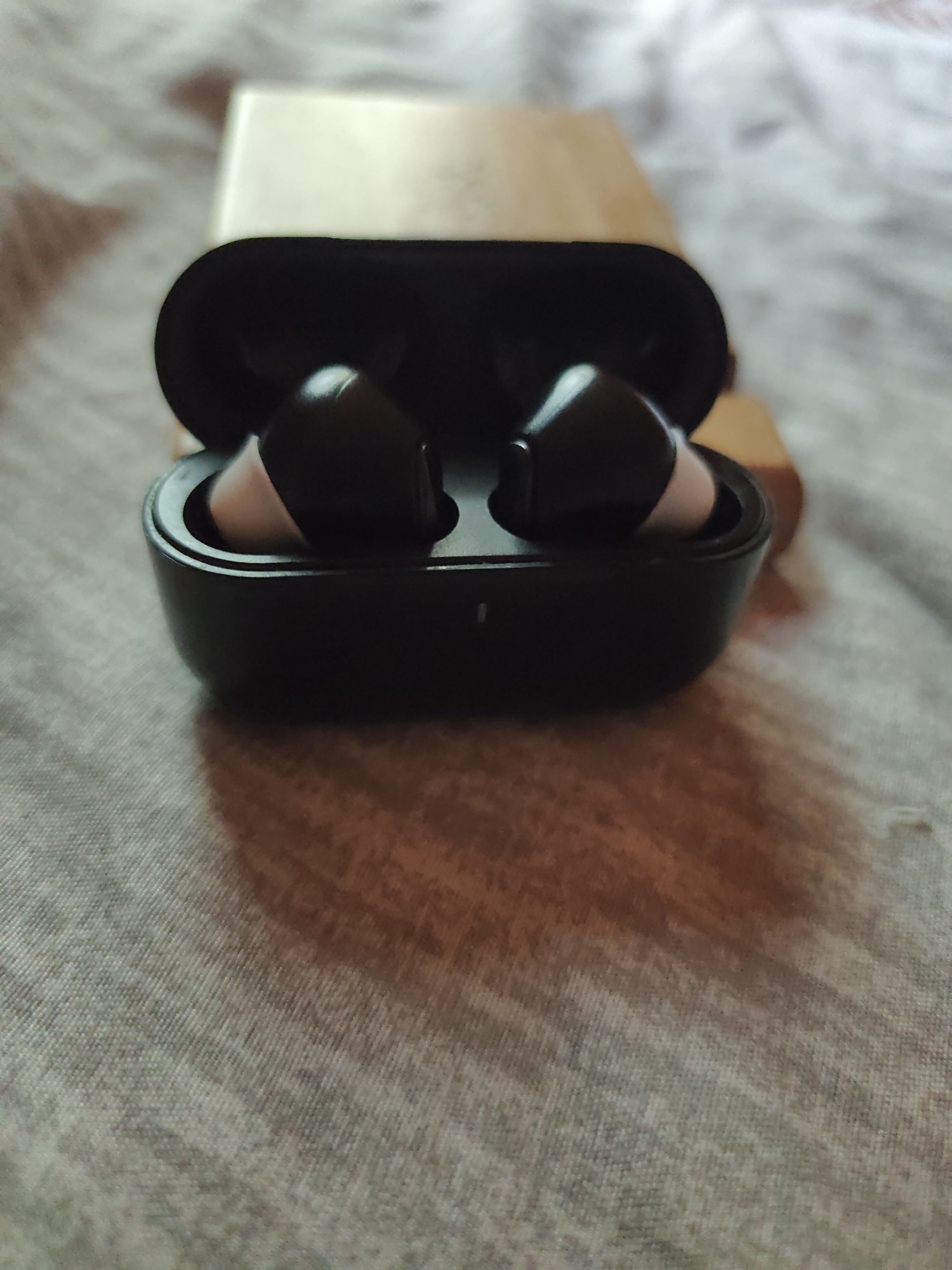 The image shows a pair of wireless earbuds in their charging case, which is open. The earbuds are black and white. The case is black and sits on top of a rectangular, light brown object, possibly a box. The background is a textured, light-colored fabric, possibly a bedspread or blanket. The location in the city of Kratovo, Russia, is not apparent from the image.