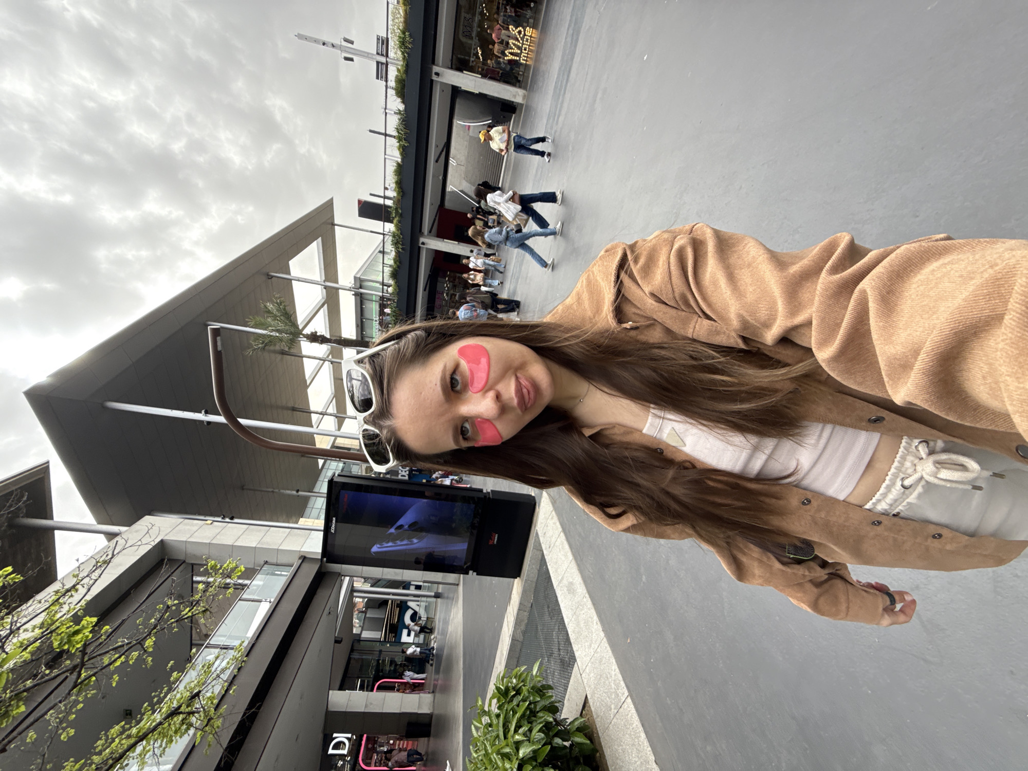 A woman is taking a selfie on a paved plaza in a city. She has long brown hair and is wearing a brown jacket, a white top, and light-colored sweatpants. She has white sunglasses perched on her head, and pink patches are on her face, below her eyes. In the background are other people, storefronts, trees, and modern architecture with a grey overcast sky. A digital billboard is nearby displaying a spaceship. The location is likely a modern shopping area with outdoor space. Although the precise location cannot be determined with certainty from the visual information, it does not look like Barcelona, Spain.