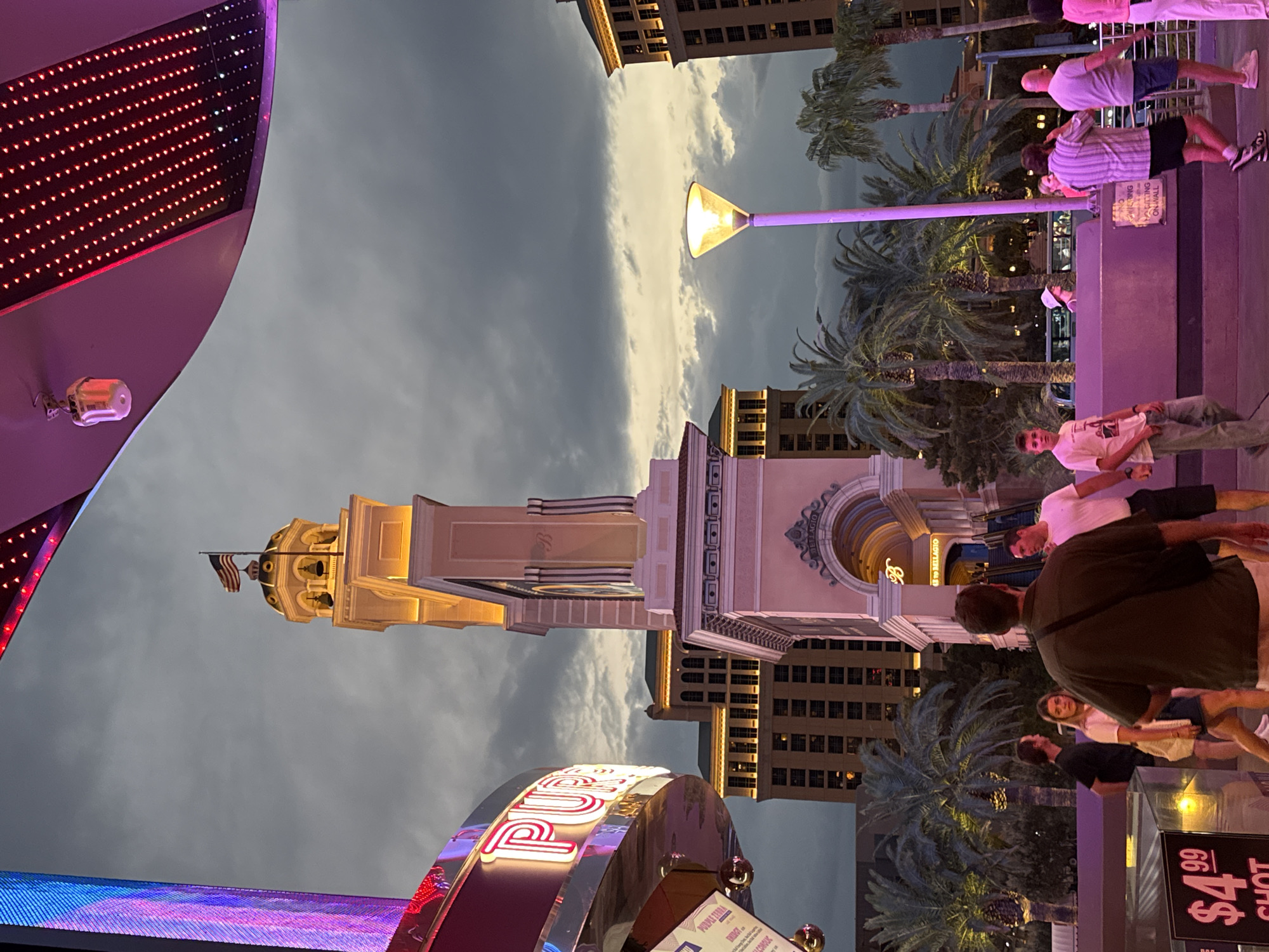 This is a medium shot of a street scene at dusk in Paradise, Nevada, USA. The foreground features a neon sign that reads "PURPLE," a street lamp with a conical shade emitting yellow light, and several people walking. The middle ground shows a tall, ornate tower with an American flag on top, a large archway, palm trees, and buildings. The background consists of a cloudy sky with hints of sunset colors. The overall atmosphere is vibrant and possibly festive, characteristic of the Las Vegas Strip.