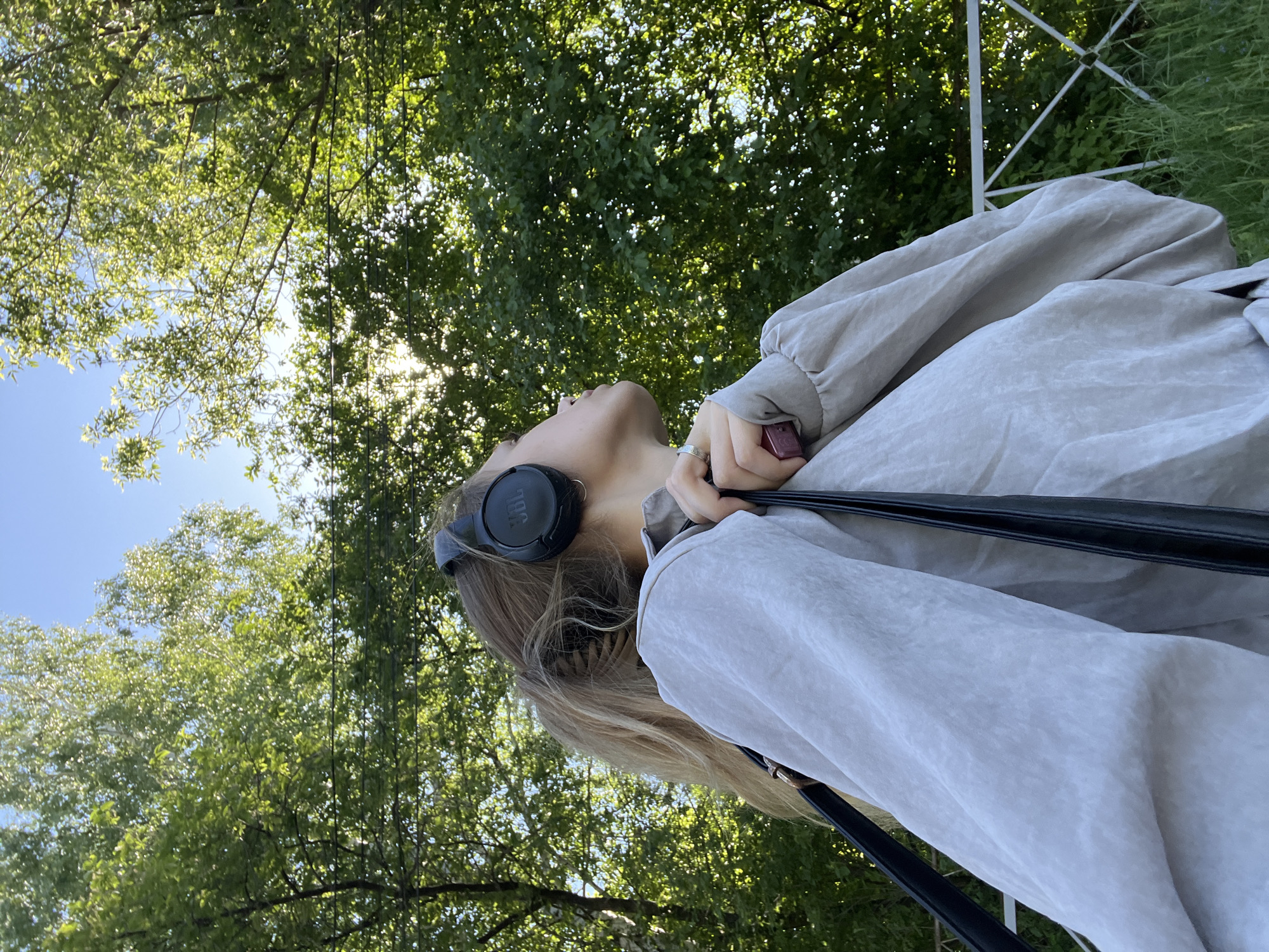 A young person with blonde hair is looking upwards. They are wearing black JBL headphones and a light gray jacket or shirt. The person is holding a small, possibly electronic, red object in their right hand. A black strap from a bag or purse crosses their body.
The background consists of green foliage from trees, with glimpses of blue sky and sunlight filtering through the leaves. To the right, there is a partially visible white metal structure, potentially part of a fence or frame.
The location is unspecified and there is no indication this image was taken in the city of Фоки, Russia.