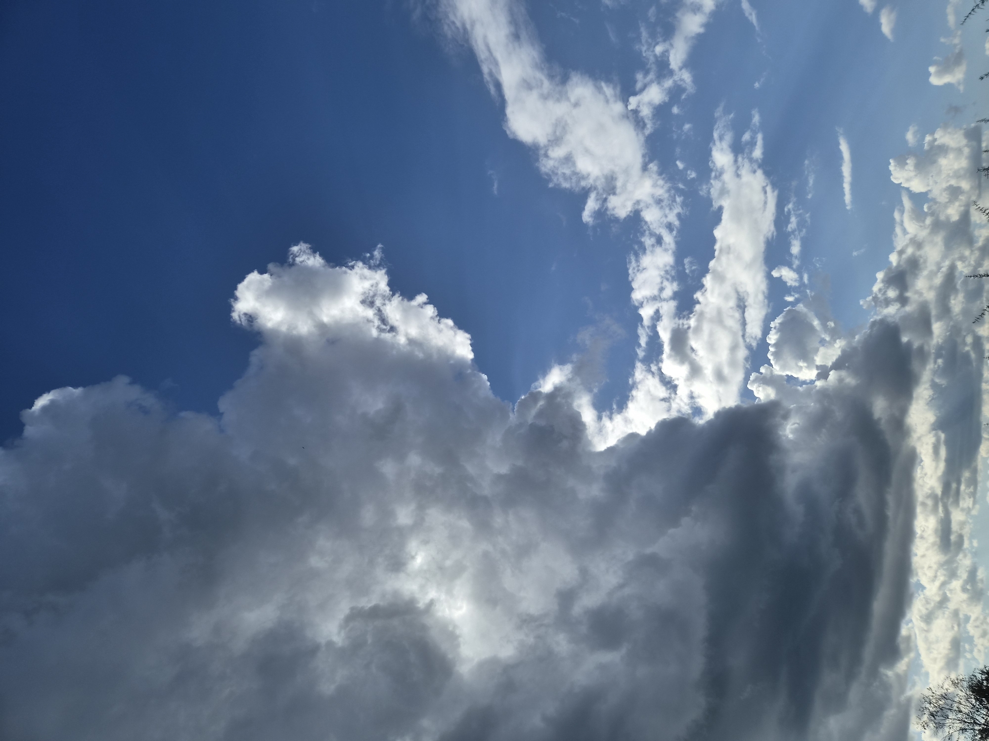 The image shows a dramatic sky with a large, dark, imposing cloud formation on the left side, transitioning into bright white clouds and a clear blue sky on the right. Sunlight filters through the edges of the dark clouds, creating a sense of depth and contrast. The lower part of the image shows hints of vegetation, possibly trees and grass, along the horizon. The sky suggests a day with changing weather conditions, possibly transitioning from stormy to clear or vice-versa. There is no indication of the city of Balyaga, Russia, or any specific human activity or urban elements within the frame.