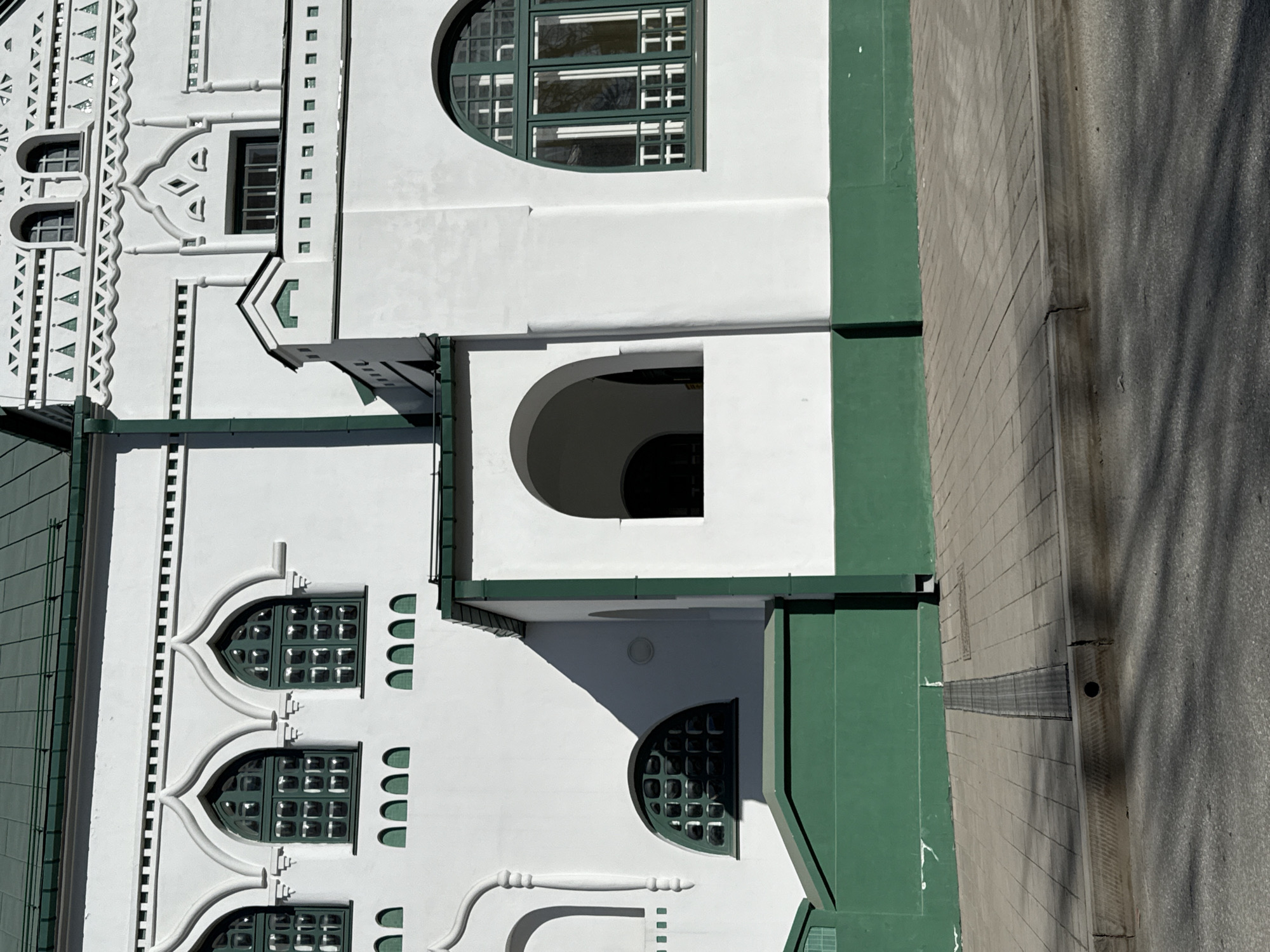 The image captures the facade of a building with distinctive architectural features, located in Perm, Russia. The building is primarily white with green accents. The architectural style includes rounded windows, arched doorways, and decorative crenellations. The building appears to be adjacent to a paved area or sidewalk. A tree is visible due to shadows on the adjacent pavement. There is no visible people or interactions.