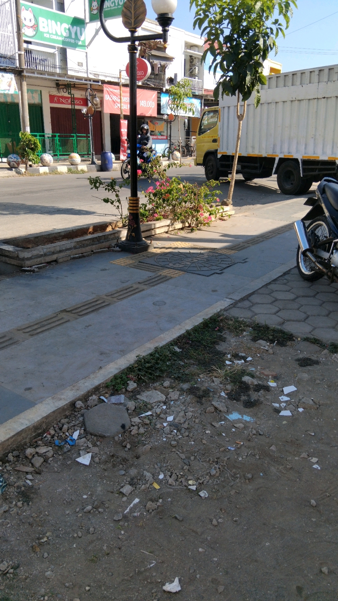 This image depicts a street scene in Batang, Indonesia. In the foreground, a patch of unkempt ground is littered with debris, including rocks, dirt, and various pieces of white and colored trash, suggesting a lack of regular maintenance. A concrete curb separates this area from a paved sidewalk. The sidewalk features tactile paving for visually impaired pedestrians and a manhole cover. Further back, a lamppost stands near a planter with pink flowers and green foliage. Across the street, buildings with storefronts are visible, including one with a sign that says "BINGYU ICE CREAM COFFEE TEA." A yellow truck is parked on the street, partially obscuring the buildings. A person wearing a helmet rides a motorcycle in the middle ground, moving away from the camera. The sky is clear and blue, indicating sunny weather.