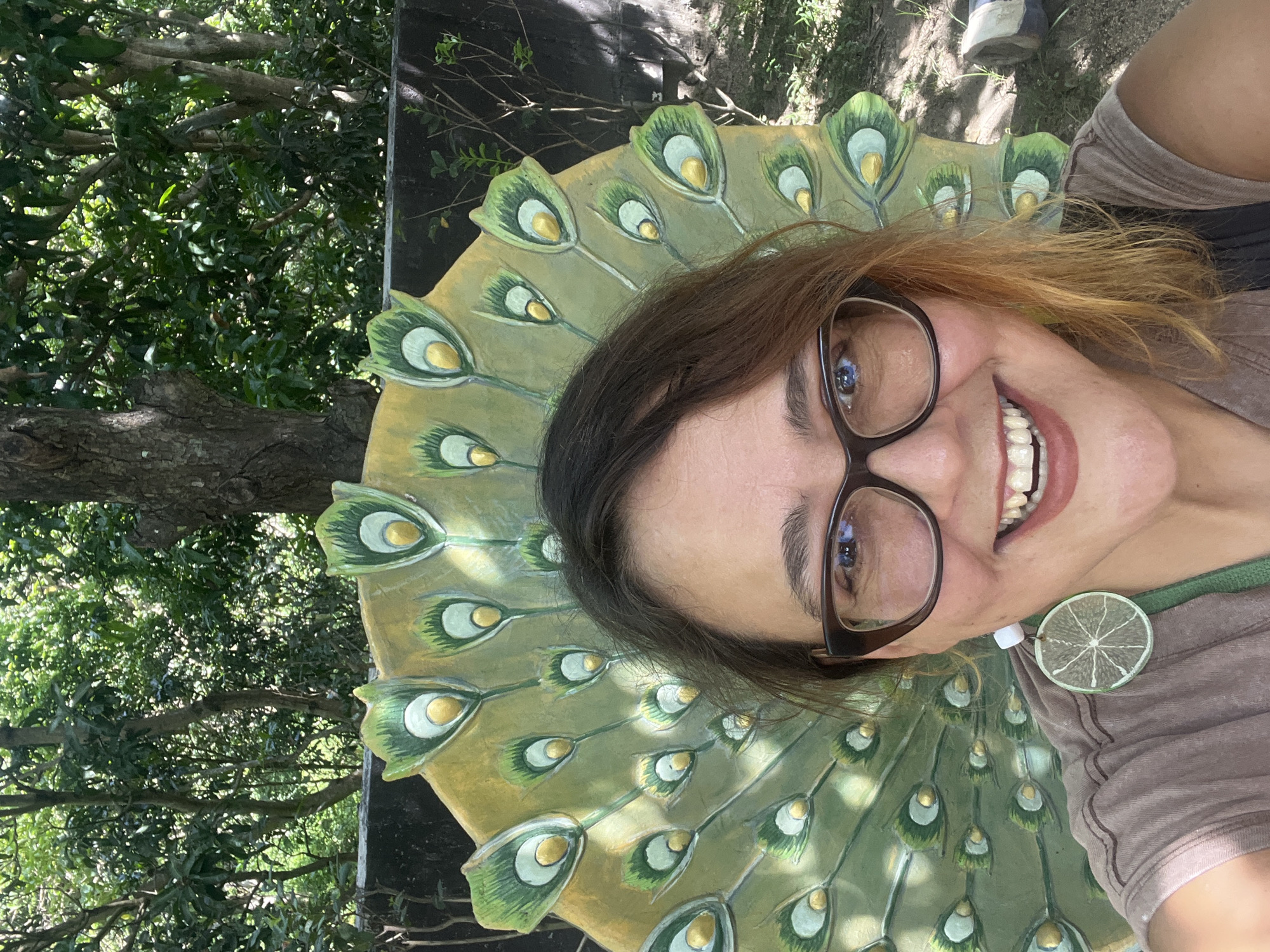 A medium-shot, eye-level, outdoor selfie features a woman with brown hair, wearing glasses and a brown t-shirt, smiling at the camera. She is positioned in front of a decorative, fan-shaped structure resembling peacock feathers, painted in various shades of green and gold. The background consists of lush green trees and foliage, suggesting a natural or park-like setting. A dark wall is visible behind the peacock structure. The woman's earrings are white and green, resembling slices of lime. The image appears to have been taken during daylight hours, with sunlight filtering through the trees. The location is identified as Nam Nha Trang Ward, Vietnam.