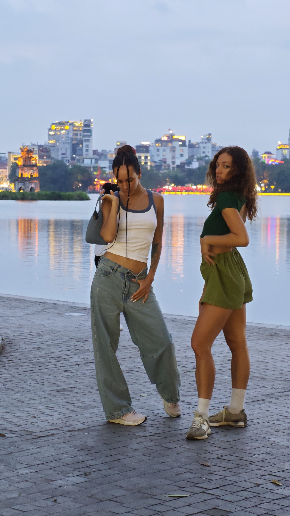 Two young women stand on a paved walkway near a body of water, with a cityscape visible in the background, likely in Hà Nội, Vietnam. The woman on the left wears a white tank top with blue straps, oversized light blue jeans, and pink sneakers. She has her hair in a bun with braids and a small blue handbag slung over her shoulder. The woman on the right wears a short-sleeved green crop top, olive green shorts, white socks, and sneakers. Both women are facing the camera. The water reflects the lights of the city in the background. The sky is overcast.