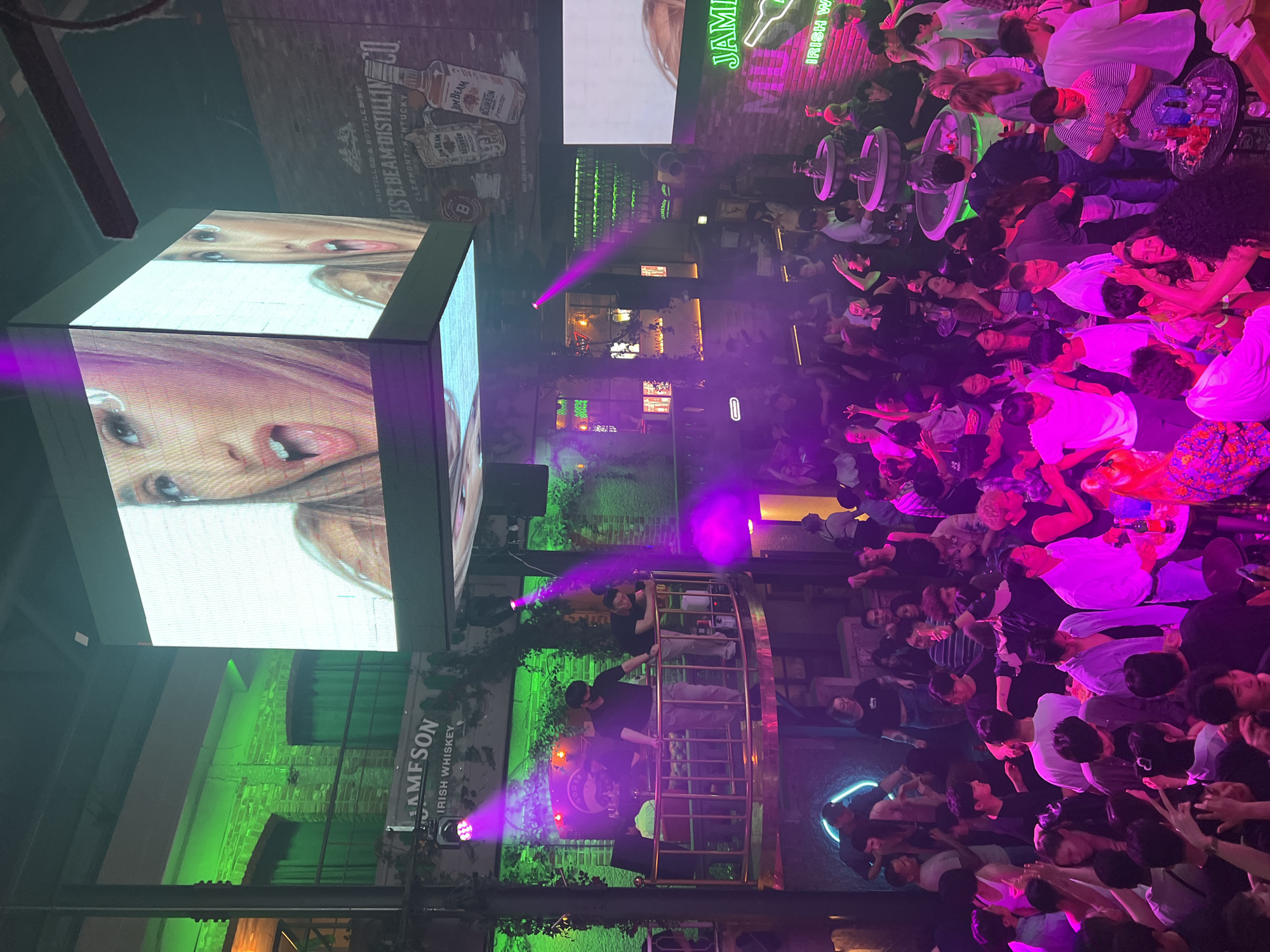 The image shows a crowded bar or nightclub scene in Seoul, South Korea. A large screen displays a close-up of a woman's face. Other branding includes Jameson Irish Whiskey and Jim Beam. Numerous people are present, densely packed and many with drinks. A metal platform structure has people on it. There is bright, colored lighting, including green and purple.