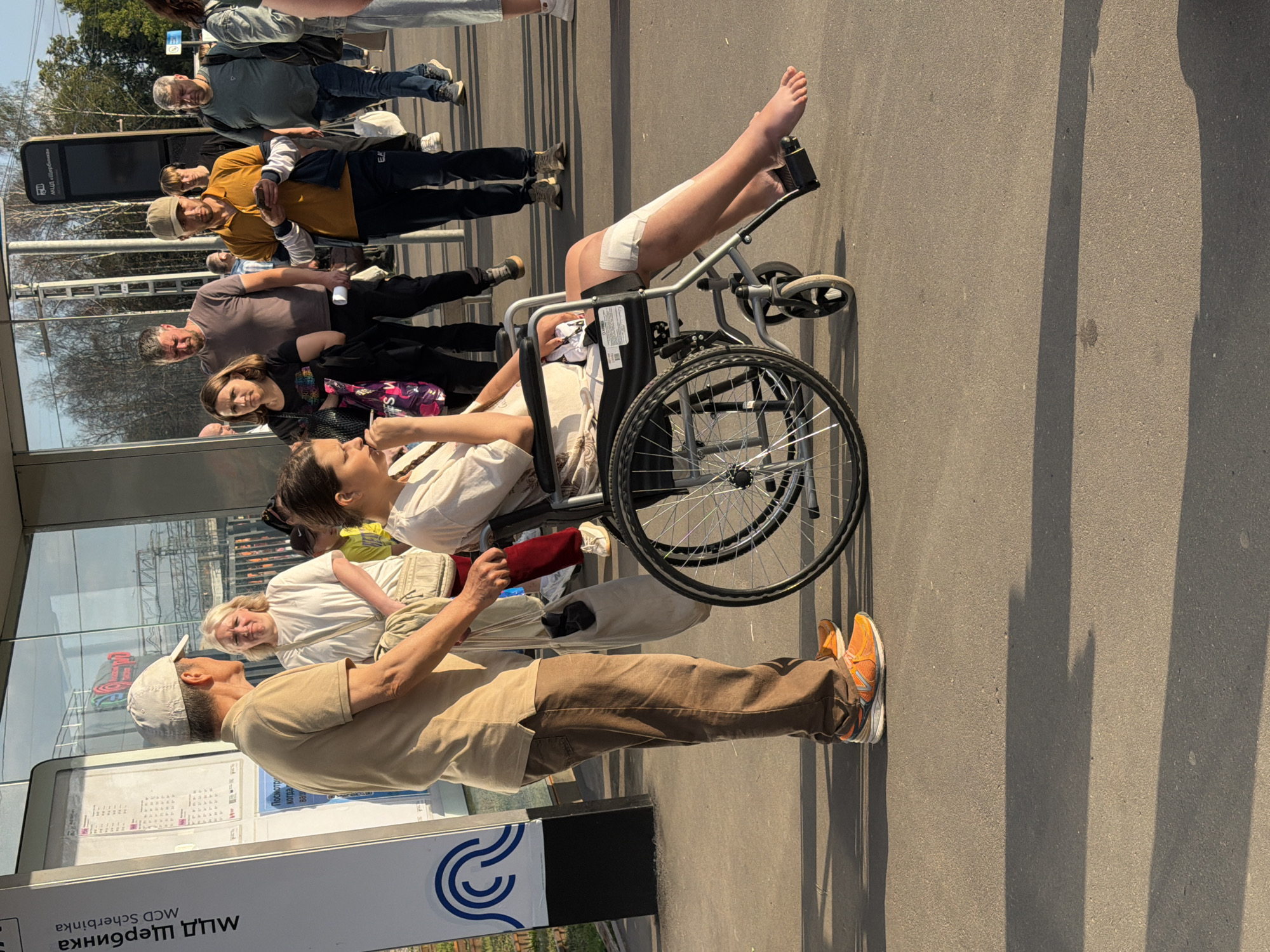 The image shows a street scene in Shcherbinka, Russia, likely near a train station given the signage. In the foreground, a person in a wheelchair is being assisted by a man. The person in the wheelchair has a bandaged leg and is barefoot on one foot. The man assisting is wearing a light brown polo shirt, brown pants, and orange athletic shoes, and is holding a bag. Other people are walking in the background, some appearing to be waiting or passing by. The environment includes a sidewalk or paved area, a bus stop or station shelter with glass panels, and trees and buildings in the distance. The lighting suggests it is daytime, possibly late afternoon due to the shadows.