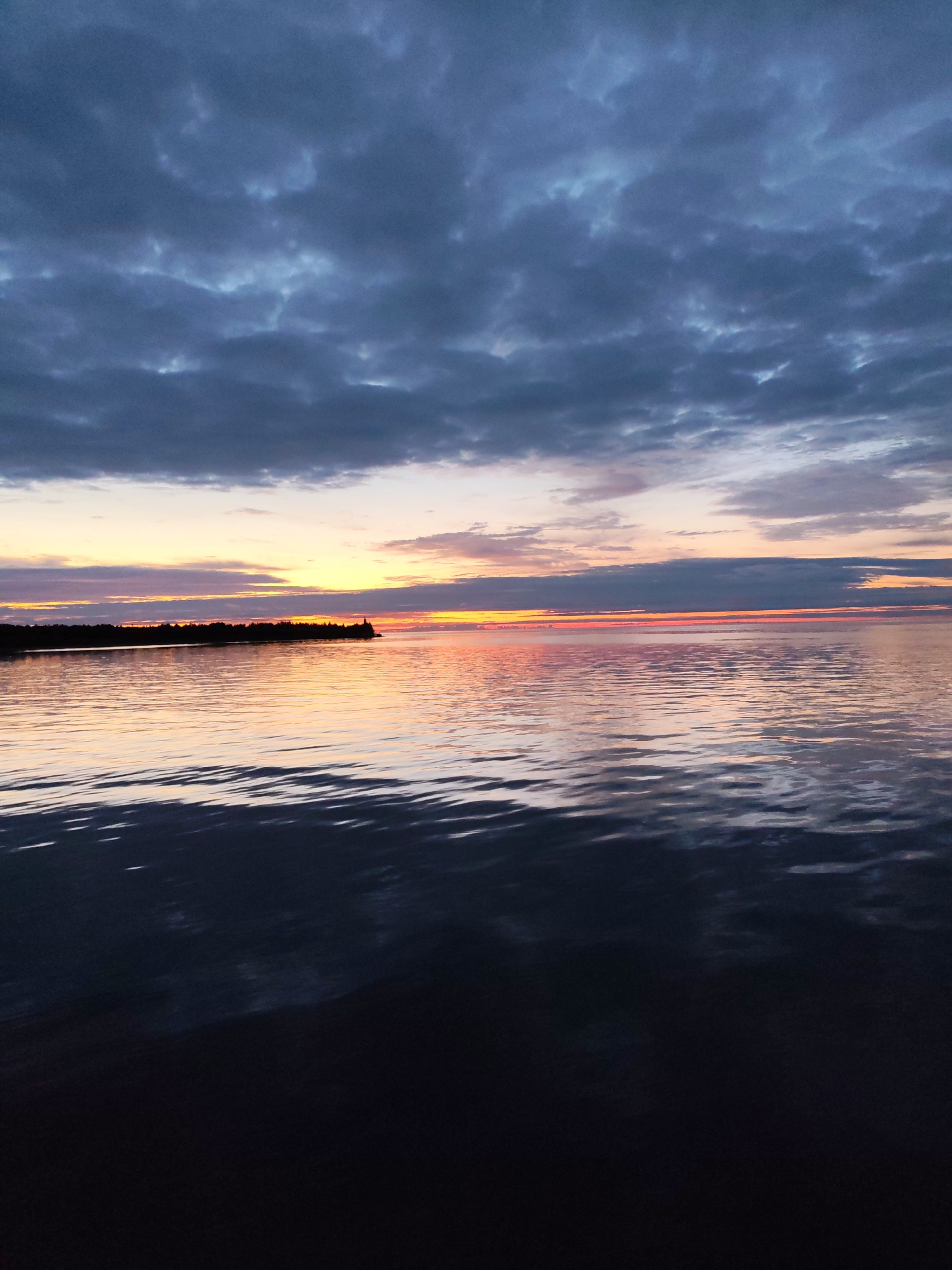 The image shows a serene sunset over a body of water, likely the Gulf of Finland near Shcheleiki, Russia. Dark, textured clouds dominate the upper portion of the frame, with hints of lighter sky peeking through in places. Below the clouds, a vibrant sunset paints the horizon with streaks of orange, yellow, and pink, reflected on the calm surface of the water. A dark, silhouetted treeline stretches across the left side of the image, punctuated by a small structure that may be a lighthouse or a building. The water in the foreground is dark and rippled, with reflections of the sky creating shimmering patterns. The overall mood is peaceful and atmospheric, capturing the beauty of a coastal sunset. There are no people or animals visible in the scene.