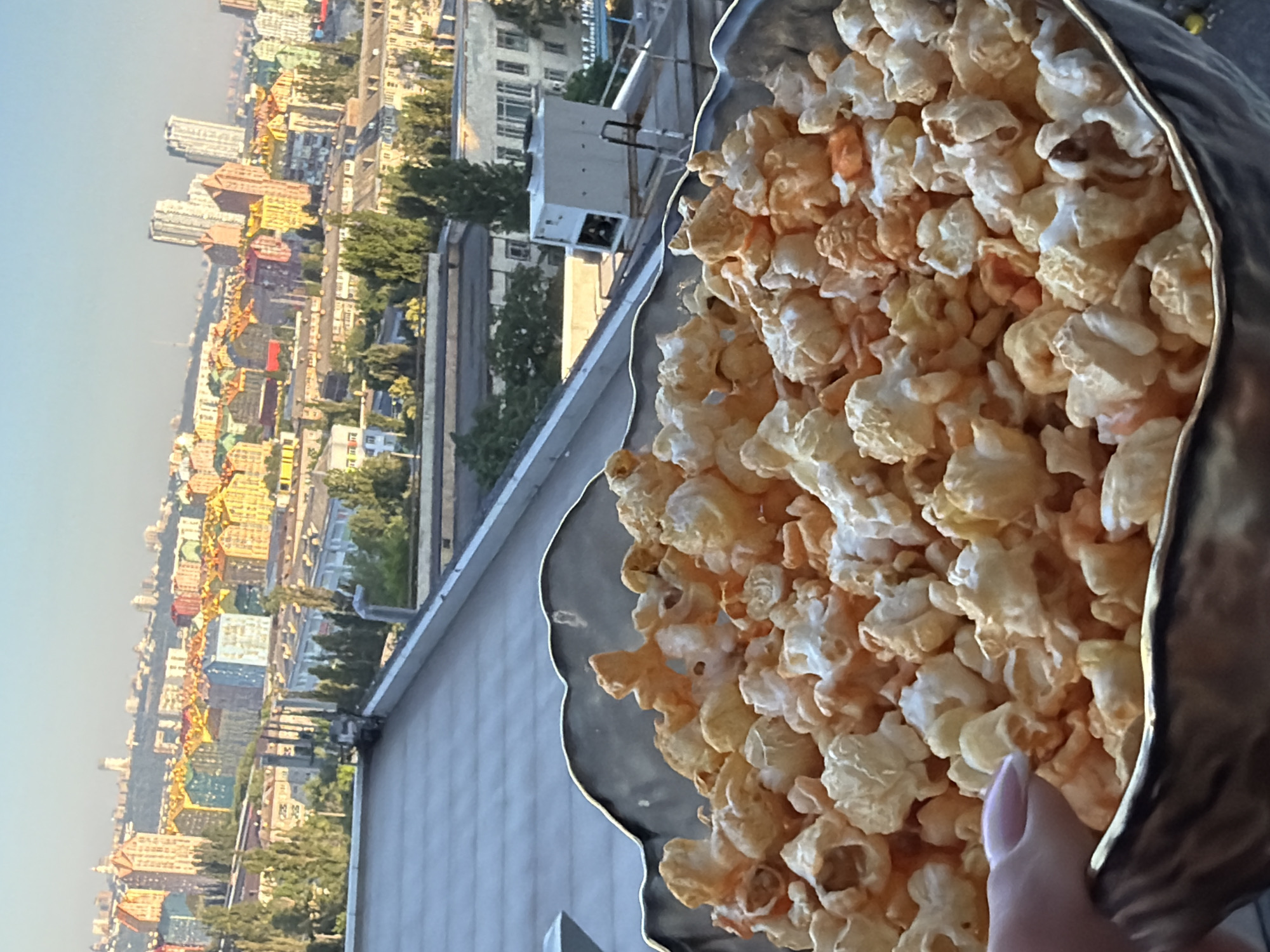 A close-up shot focuses on a bowl filled with popcorn, resting on a rooftop with a cityscape in the background. The popcorn appears to be lightly seasoned with orange and white flecks. A hand with a pink-manicured fingernail is visible on the left side of the frame, near the edge of the bowl. In the distance, the colorful residential buildings of the "Comfort Town" complex in Kyiv, Ukraine, are visible under a clear sky. The buildings are painted in a variety of bright hues, creating a vibrant contrast with the more muted tones of the rooftop and surrounding structures. The scene is captured during daylight, likely late afternoon, given the warm lighting.