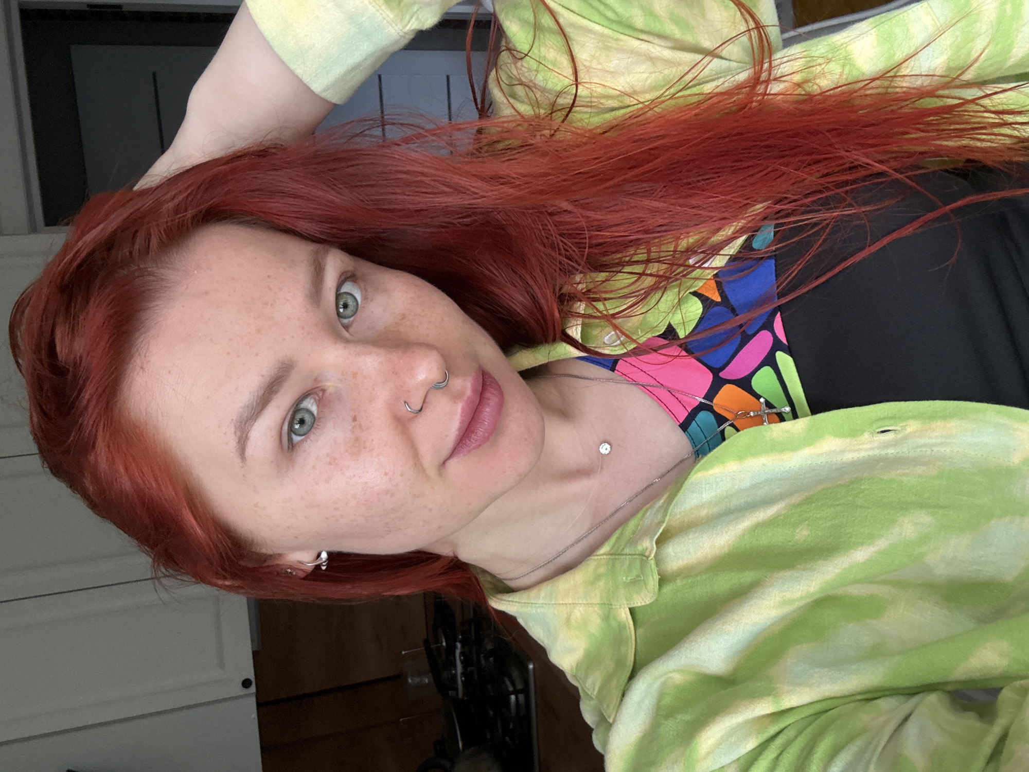 A selfie of a woman with red hair, green eyes, and freckles, wearing a tie-dye shirt, a colorful top underneath, and jewelry (earrings, nose ring, necklace, collarbone piercing). The background is nondescript; part of a white door or cabinet is visible on the left. No information suggests the location is Rostov-on-Don.