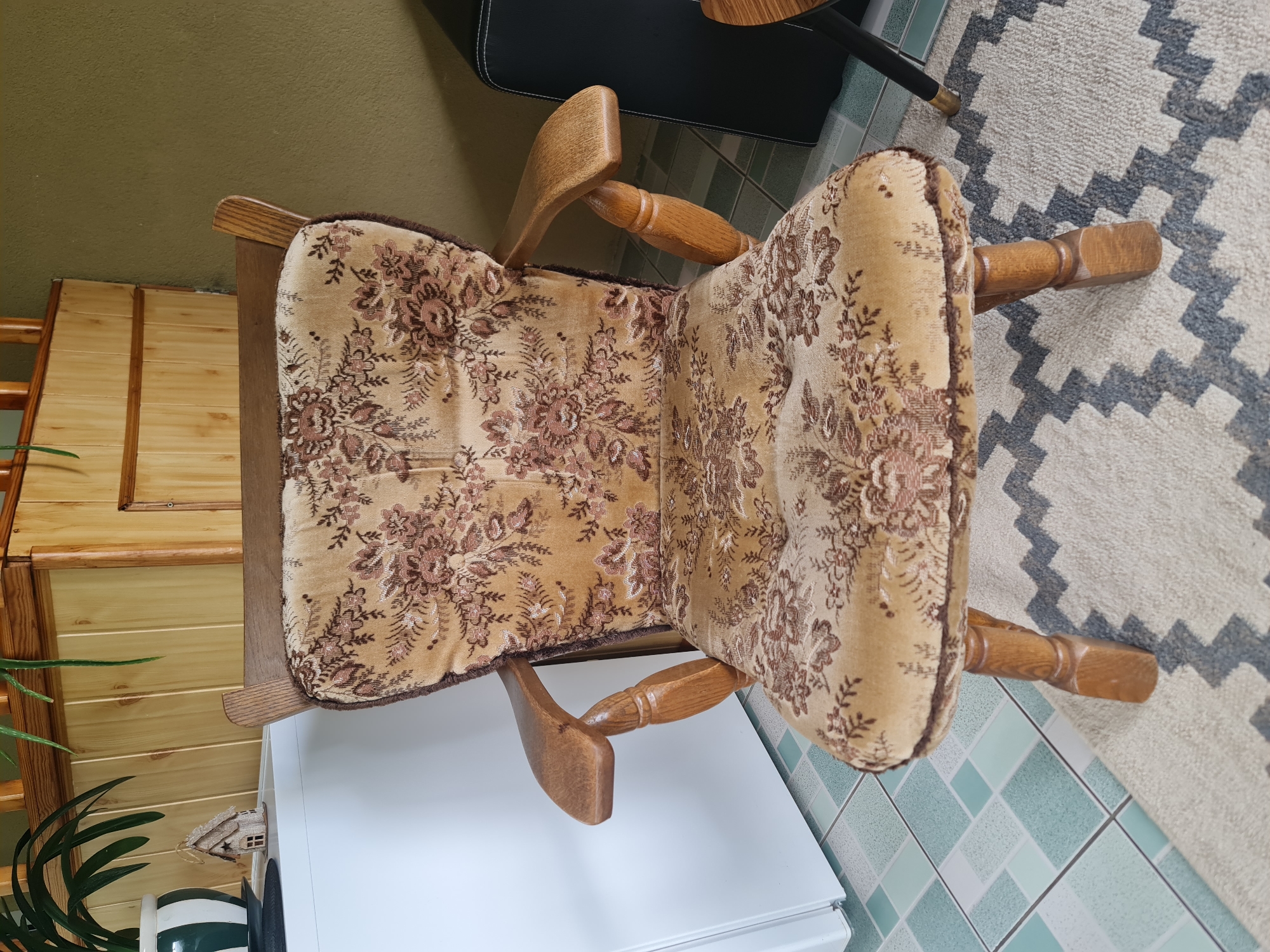 An antique wooden armchair with a floral upholstered seat and back is positioned on a patterned rug. The chair has carved wooden arms and legs. Behind the chair is a light-colored cabinet made of wood planks, and to the left is a white appliance, possibly a refrigerator. The floor is tiled with a geometric pattern of light blue and white squares. In the upper left corner, a plant in a green and white pot is visible, along with a small decorative house. The wall behind the chair is painted a muted olive green. The scene appears to be indoors, in what might be a residential setting in Molėtai, Lithuania. There are no people or animals present.