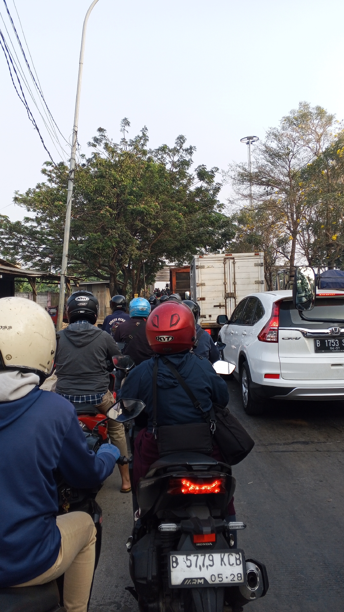 The image depicts a traffic jam on a street in Bekasi, Indonesia. In the foreground, several motorcyclists are seen from behind, wearing helmets of various colors including beige, black, blue, and red. The motorcyclists are seated on their vehicles, indicating they are stationary or moving slowly. One motorcyclist wears a blue hooded jacket and beige pants, while another wears a grey hooded jacket and plaid pants.
Behind the motorcycles, a white truck is visible, suggesting it is part of the congestion. To the right of the truck, a white Honda CR-V SUV is also present in the traffic. The street is paved, and there are trees lining the sides of the road, with some foliage obscuring the sky. Utility poles and wires are visible in the upper left portion of the image, indicating urban infrastructure. The overall scene suggests a typical urban traffic condition during daylight hours.