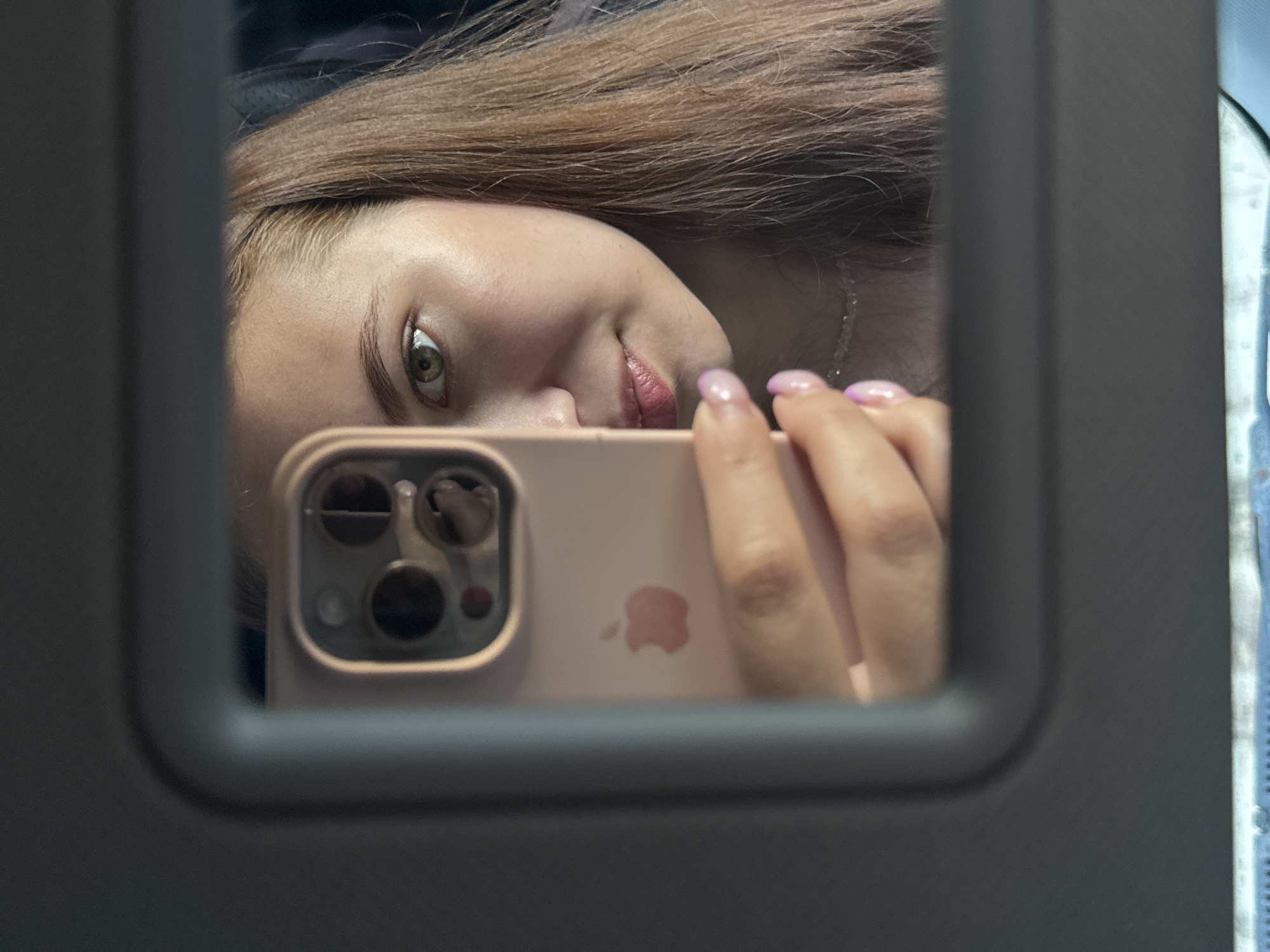 A young woman with long, auburn hair takes a selfie in a car's vanity mirror. She is holding a pink iPhone in her left hand, and her face is partially obscured by the phone. Her right eye is visible, and she is looking directly at the camera. Her lips are slightly parted, and she is wearing a light-colored necklace. The background is dark and out of focus, suggesting the interior of a vehicle.