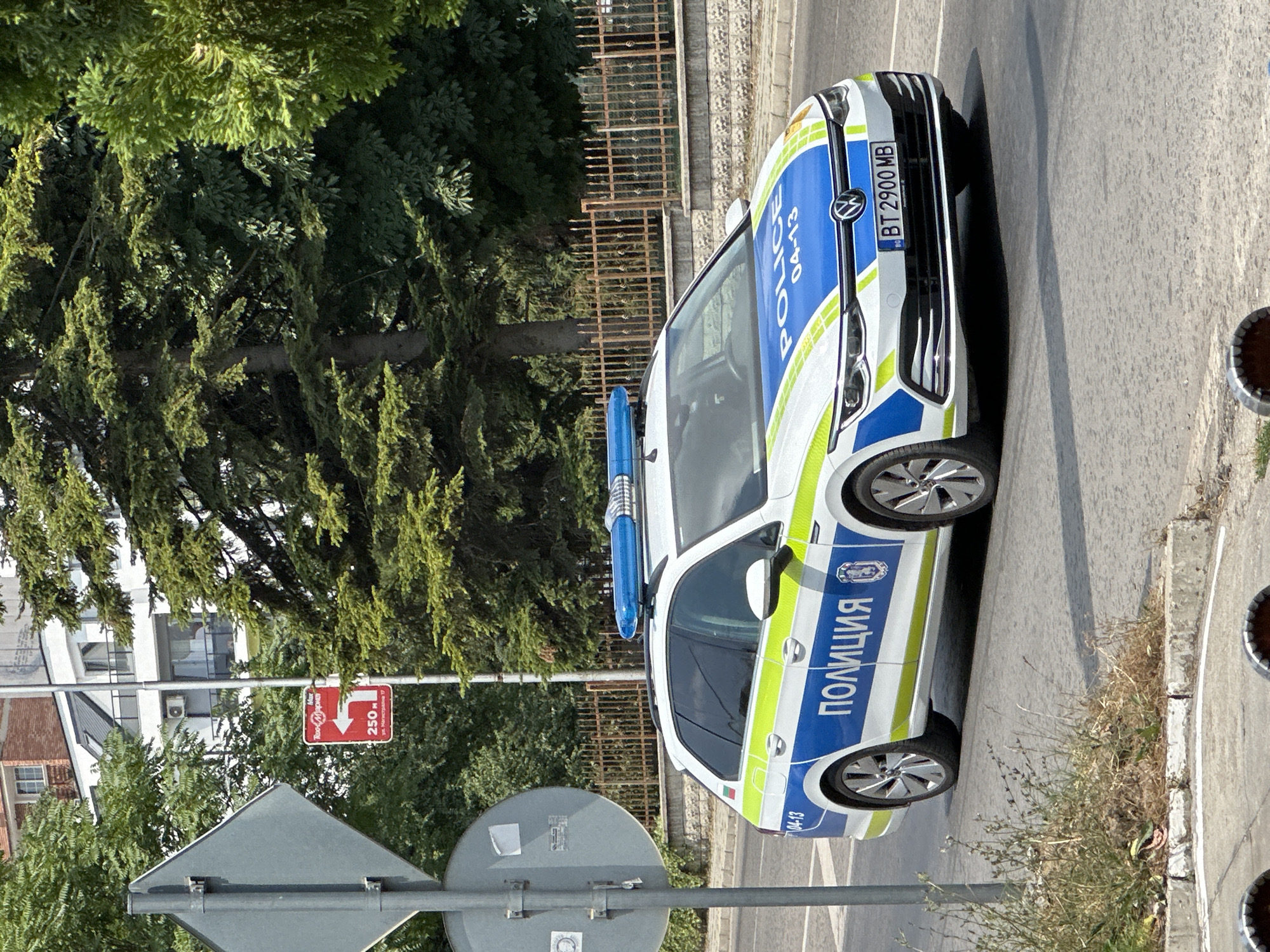 A police car is parked on a street in Veliko Tarnovo, Bulgaria. The car is white with blue and yellow markings, and has the word "POLICE" written on the side in English and "ПОЛИЦИЯ" in Cyrillic. The license plate reads "BT 2900 MB". The scene appears to be during the daytime, under clear skies, given the shadows cast by the car and trees. Green trees and a metal fence are visible in the background, as are parts of buildings and street signs. One sign shows a red arrow pointing left with the text "Max 20 km/h" and "250 m", indicating a speed limit and distance to a turn. The overall impression is of a typical day in a European city. No people are visible in the frame.