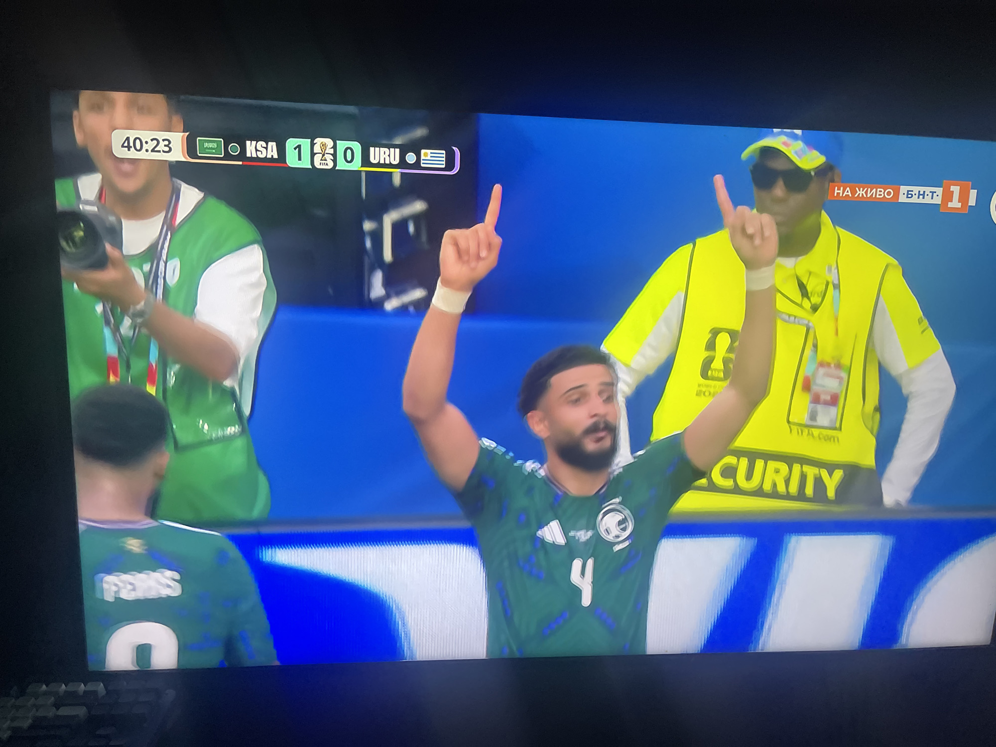 The image captures a moment from a soccer match, displayed on a television screen, showing the scoreline "40:23 KSA 1 0 URU". In the foreground of the screen, a male soccer player wearing a green jersey with blue patterns and the number "4" is celebrating with his arm raised, pointing upwards. To the right of the player, a security guard in a bright yellow vest and sunglasses is also visible, with his arm raised in a similar gesture. Behind them, the stadium's blue backdrop and white seating are partially visible. The broadcast overlay includes Bulgarian text and a logo for "BHT 1". The image is framed by the black bezel of the television.