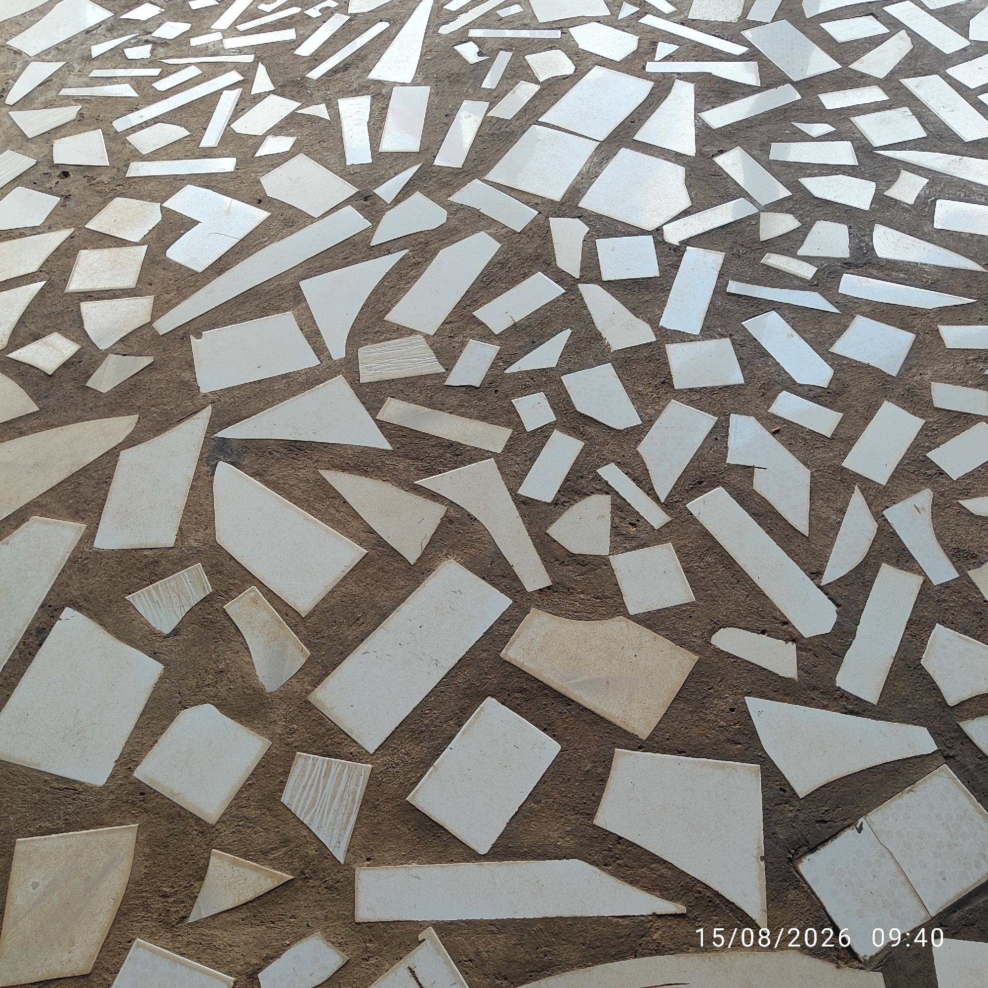 The image shows a mosaic pattern composed of broken white tiles set into a dark, coarse aggregate concrete ground surface. The tiles are irregularly shaped and vary in size, creating a fragmented, abstract design. The ground is textured and appears to be outdoors, possibly a pathway or an outdoor floor. The lighting suggests it is daytime. A timestamp in the lower right corner reads "15/08/2026 09:40", indicating the date and time the photograph was taken. The location is identified as Buding, Indonesia. There are no people or animals visible in the image.