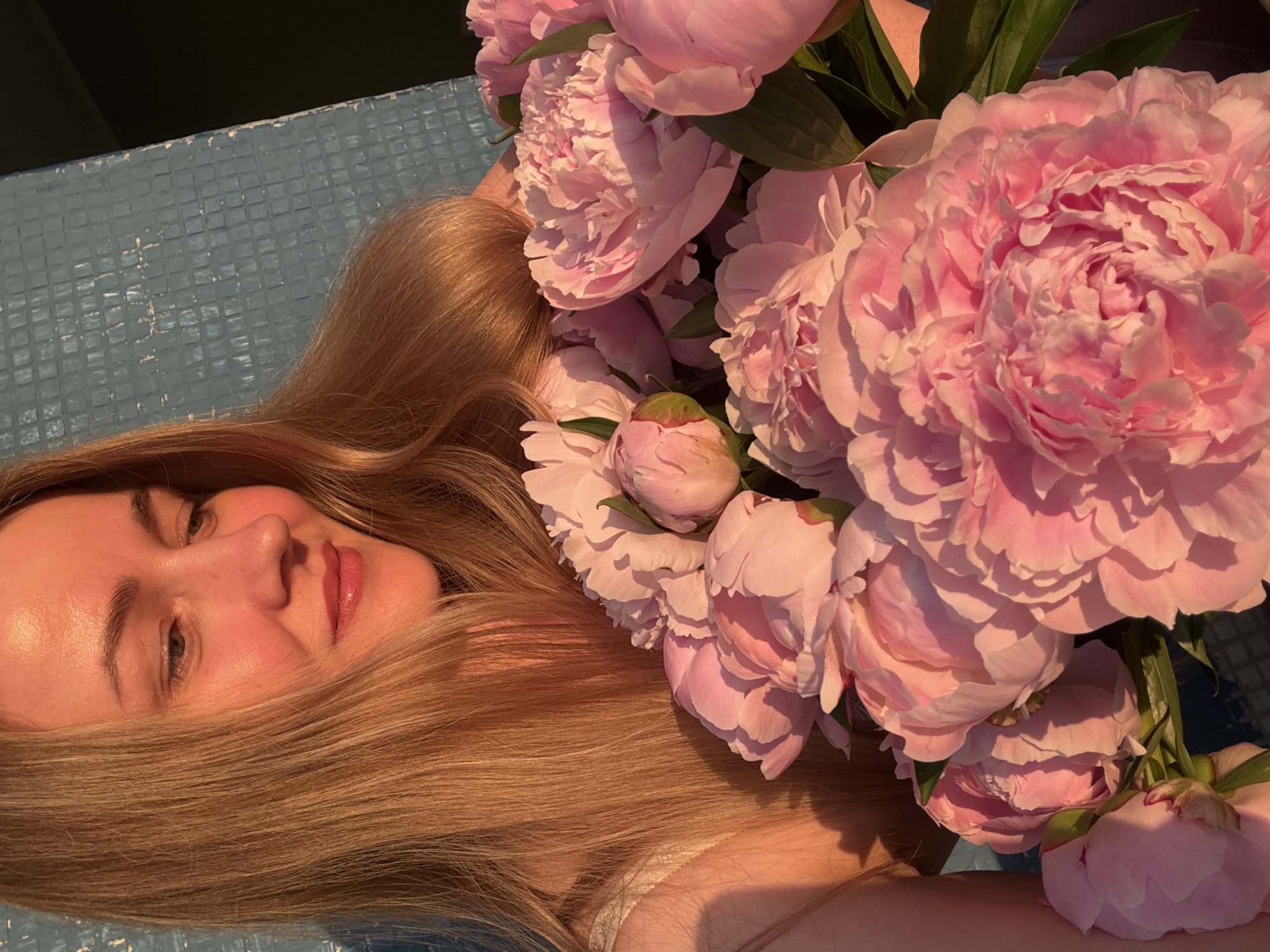 In Chaikovsky, Russia, a young woman with blonde hair is illuminated by the warm, golden light of the setting sun. She is holding a large bouquet of pink peonies, their petals soft and full. Her gaze is directed downwards, a gentle smile gracing her lips, as if lost in thought or admiring the flowers. Behind her, a textured blue wall, possibly a building exterior, serves as a backdrop, its surface marked by a grid of small squares. The scene evokes a sense of tranquility and natural beauty, bathed in the soft glow of twilight.