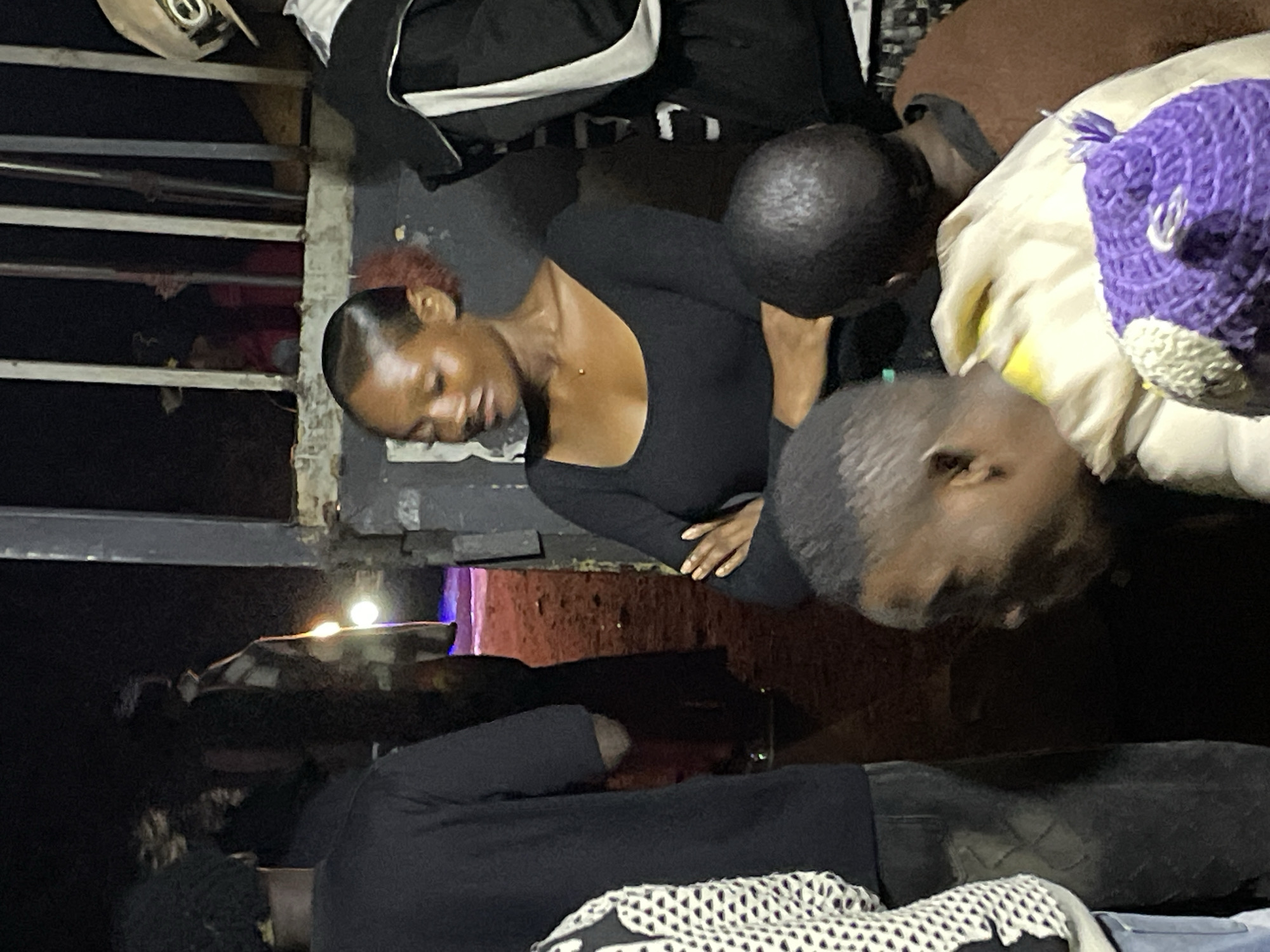 In Wumat, Nigeria, a group of individuals are gathered near an opening in a fence. A young woman with her hair pulled back and a few red strands visible is looking down and to her right, her arms crossed. She is wearing a black, long-sleeved shirt and a small necklace. To her left, a person wearing a black shirt and jeans is partially visible from the back. To the right of the woman, another person wearing a black jacket with white stripes is standing. In the foreground, two individuals are partially visible. One is wearing a light-colored jacket over a brown garment and a purple crocheted hat with white and yellow accents. The other person is seen from the side and is wearing a brown garment. The background appears to be a dirt path with some lights visible in the distance. The scene has a dim, nighttime ambiance, possibly indicating an outdoor event or gathering.