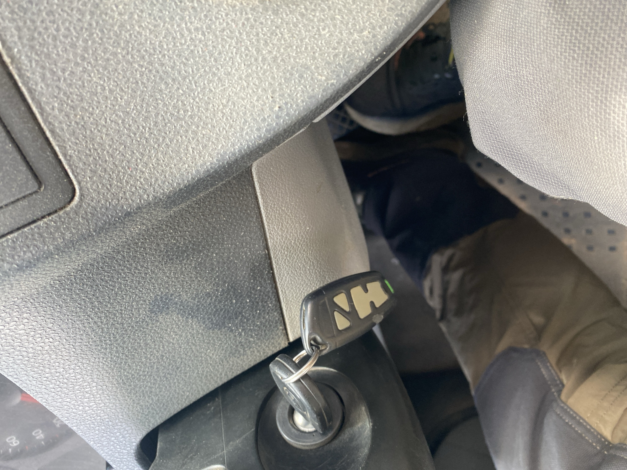 A close-up, eye-level shot shows a car's ignition and a key fob. The ignition is on the left side of the frame, with a key inserted. Attached to the key ring is a black key fob with several buttons. The key fob has a green indicator light at the bottom. The car's steering column and dashboard are visible in the background, appearing dusty. The lower portion of the frame shows a person's legs, clad in pants and shoes, indicating the perspective is from inside the car. The scene is indoors, likely within a vehicle parked in an urban setting.