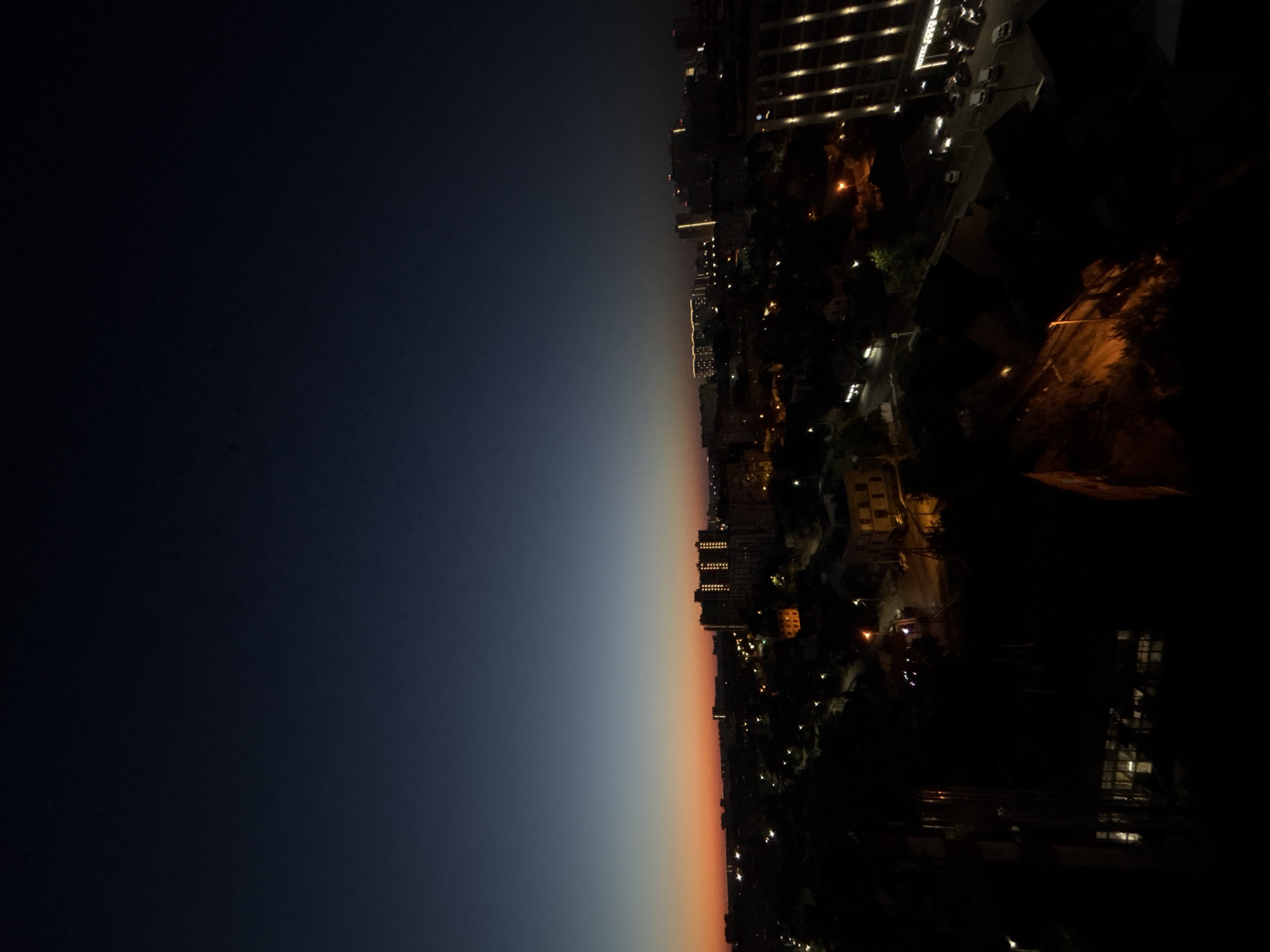The image captures a dusky cityscape, likely at dawn or dusk, with the sky transitioning from a deep navy blue at the top to a soft orange hue near the horizon. Below the sky is a dark panorama of buildings and city lights. In the mid-ground, several apartment buildings are visible, some illuminated with strings of warm yellow lights, particularly a large building on the right with vertical rows of lights. Scattered streetlights and lights from windows dot the urban landscape, creating a sense of activity within the dimness. The foreground is very dark, with the silhouettes of trees and what appear to be other buildings obscuring much of the detail. The overall impression is of a developing city scene as natural light fades or begins to emerge. The presence of tall apartment blocks and a general urban layout are consistent with a location in Krasnodar, Russia.