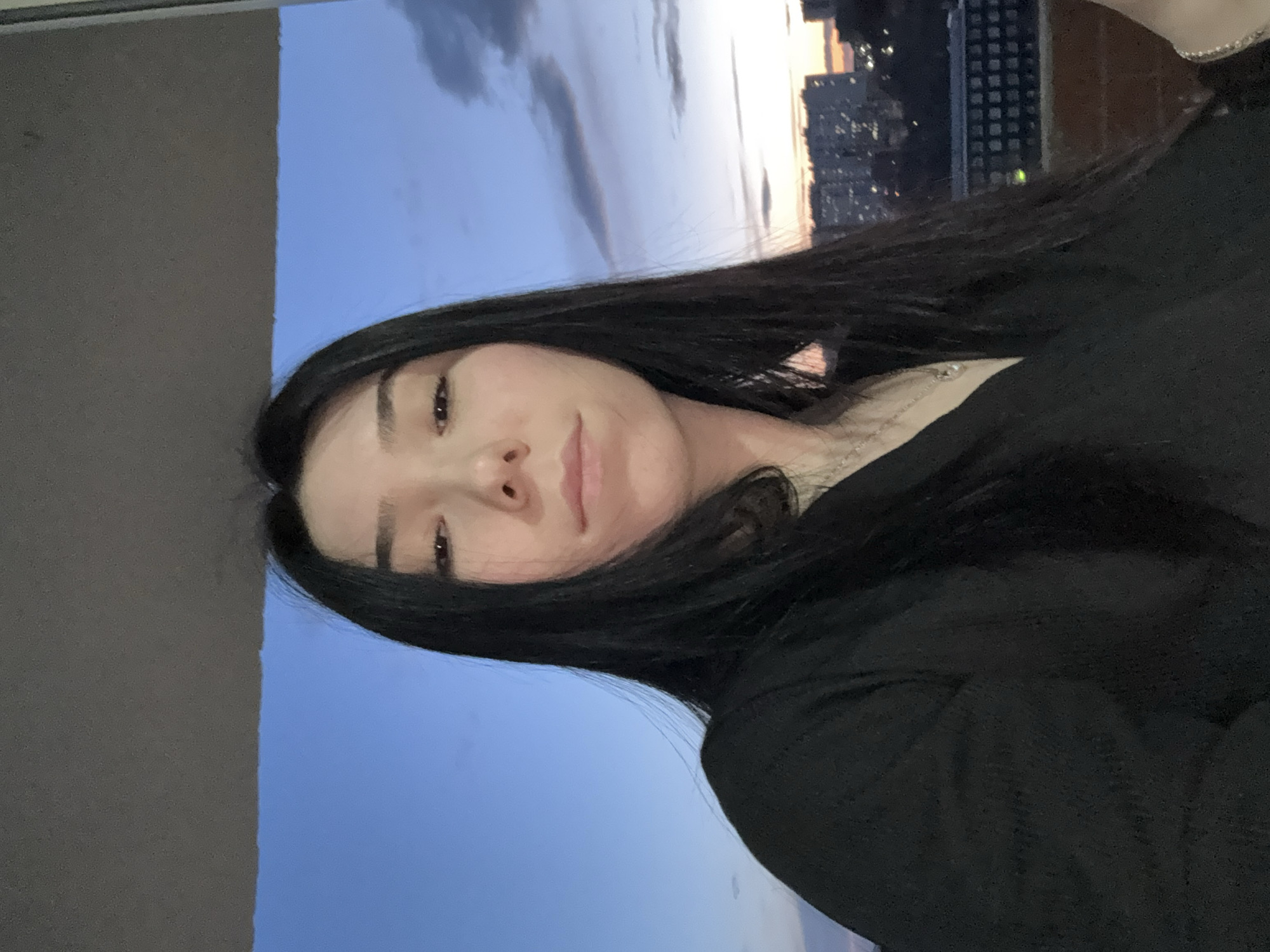 The image is a portrait-style photograph, rotated 90 degrees counter-clockwise from a standard upright orientation. The central subject is a young woman with long, dark hair, fair skin, and a serene expression, looking directly at the camera with a subtle smile. She appears to be wearing a dark, possibly black, long-sleeved top, and a delicate silver-colored necklace with a small pendant is visible around her neck.
Behind her, filling the background, is an urban landscape at dusk in Izhevsk, Russia. The sky transitions from a deep, clear blue to lighter orange and yellow hues near the horizon, illuminated by the setting sun. Some faint clouds are scattered across the sky. Below the horizon, the silhouette of a city skyline with various buildings, some displaying illuminated windows, indicates an active urban environment. A dark, plain wall or interior frame occupies the far left edge of the image as presented. The time of day is clearly twilight, characterized by the dramatic sky colors and city lights. The overall mood is calm and contemplative. No text is visible in the media file.