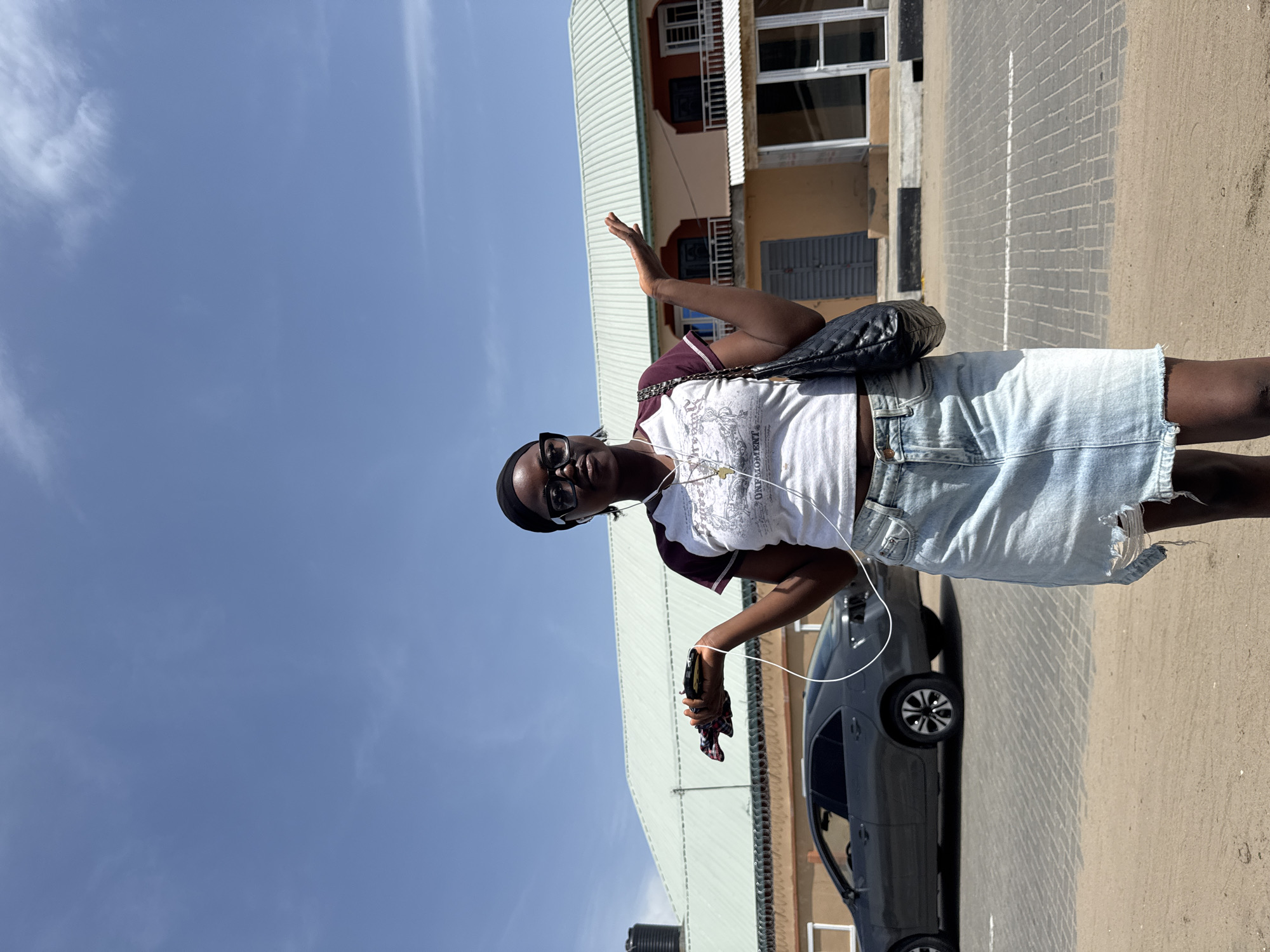 A woman stands on a paved surface on a sunny day in what appears to be Badagry, Nigeria. She has dark skin, is wearing a white t-shirt, a light-colored denim skirt with frayed edges, glasses, and a head covering. Headphones hang around her neck. She holds a phone or small device in one hand and has the other hand raised in a wave. A black purse or bag is slung over her shoulder. Behind her, a dark-colored car is parked next to a building with a light-colored corrugated metal roof. The building appears to have multiple levels with some windows and doors visible. The sky is blue with some clouds. The setting suggests an urban or semi-urban environment. The woman appears to be interacting with someone or something outside the frame.