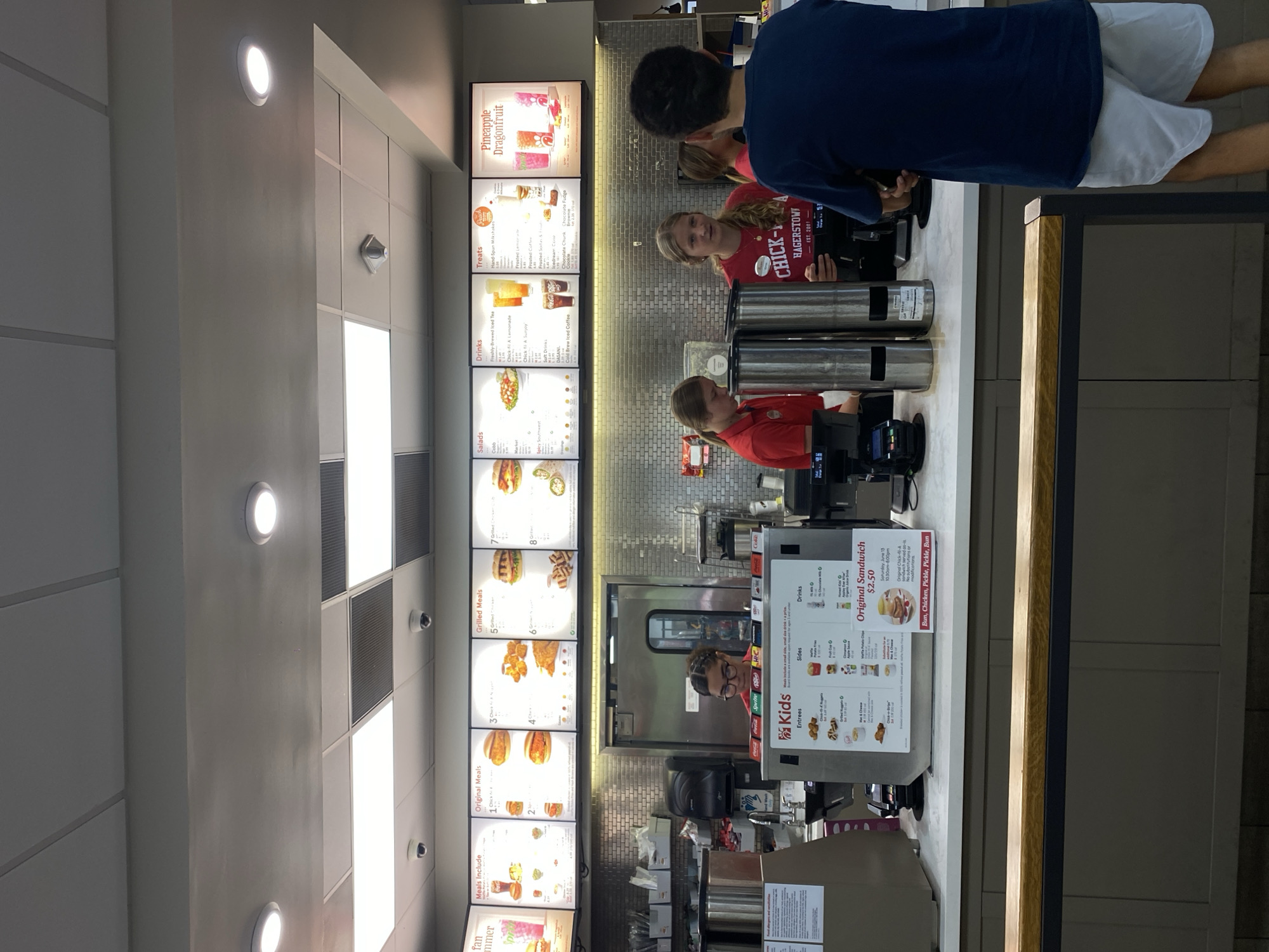 The image depicts the interior of a Chick-fil-A restaurant, likely in Hagerstown, Maryland, United States of America, based on the visible branding on an employee's shirt. The scene is a counter area where customers interact with staff.
In the foreground, a young male wearing a dark blue t-shirt and light-colored shorts stands facing away from the camera, looking towards the counter. Behind him, a counter separates the customer area from the food preparation space. On the counter, there are various items including a credit card reader, a menu board displaying "Kids" meals, entrees, sides, and drinks, and a "Original Sandwich $2.50" sign. There are also two tall, stainless steel beverage dispensers.
Behind the counter, three young female employees are visible. One, in a red shirt, is interacting with a customer or colleague. Another, also in a red shirt with "CHICK-FIL-A HAGERSTOWN EST. 2001" printed on it, is looking towards the camera and appears to be a cashier or server. A third employee, in a red shirt, is partially visible further back. The wall behind the counter is covered with a textured, light-colored tiled backsplash. Above the counter, a menu board displays various food and drink options, categorized by "Meals Include," "Original Meals," "Grilled Meals," "Salads," "Drinks," "Treats," and a special offering of "Pineapple Dragonfruit." The ceiling features recessed lighting and acoustic tiles. A security camera is visible mounted on the ceiling. The overall environment suggests a typical fast-food restaurant setting during operational hours.