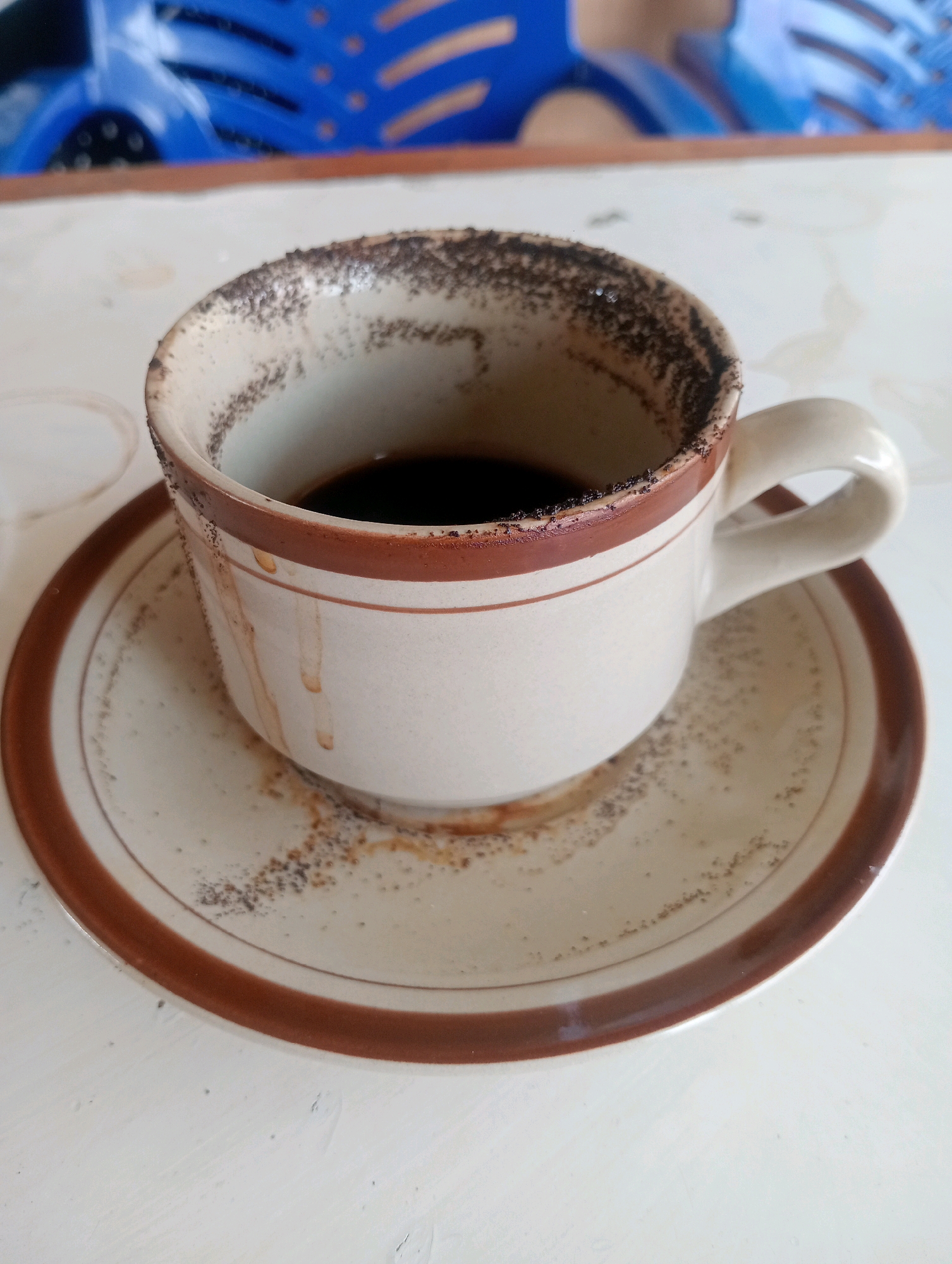 A close-up shot captures a ceramic coffee cup and saucer on a white table. The cup, white with brown trim, contains dark coffee and has coffee grounds clinging to its rim and the inside of the cup. The saucer, also white with brown trim, has a scattering of coffee grounds on its surface, and two brown streaks run down the side of the cup. In the background, a blurry blue plastic chair is visible. The image appears to be taken in Plosojenar, Indonesia.