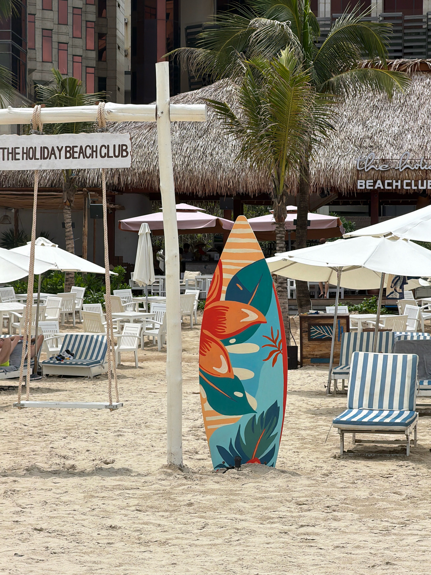 The image depicts "The Holiday Beach Club" located on the beach in Đà Nẵng, Vietnam. The scene features a sandy beach with numerous white lounge chairs and tables set up, some with umbrellas. In the foreground, a surfboard with a colorful tropical design stands upright in the sand. To the left, a wooden swing hangs from a white post. Behind the beach furniture, a thatched-roof building with the club's name visible can be seen. Palm trees are scattered throughout the scene, and in the background, modern buildings rise above the beach club. A few people are visible sitting on chairs or walking in the distance. The overall environment suggests a relaxed beach resort atmosphere.